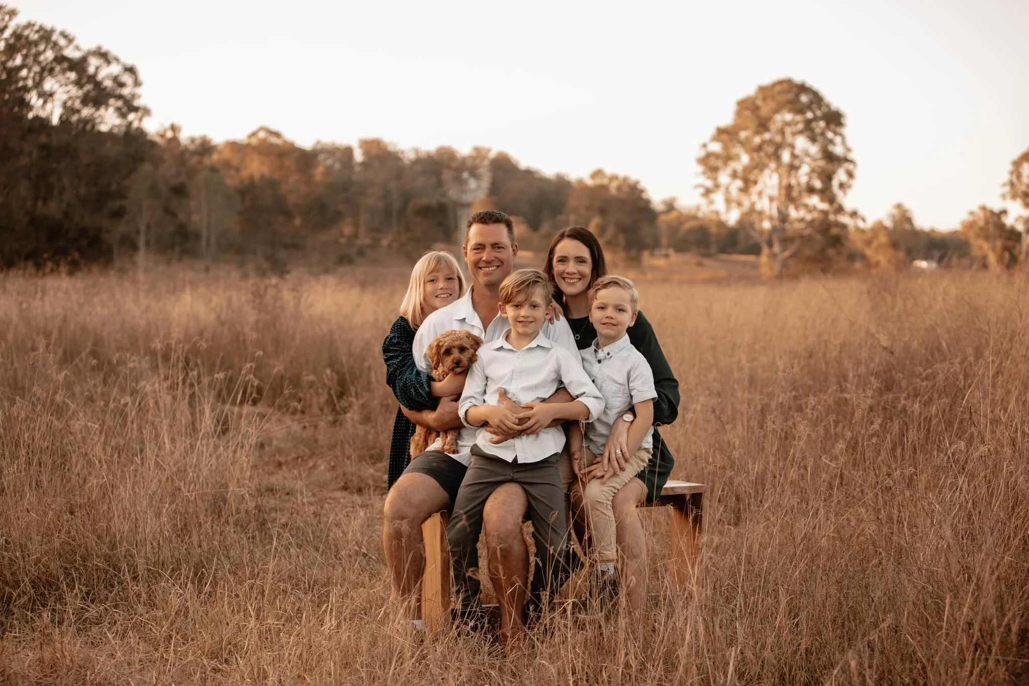 A family of five with two children and a dog sitting on a wooden bench in a field of tall, dried grass during sunset.