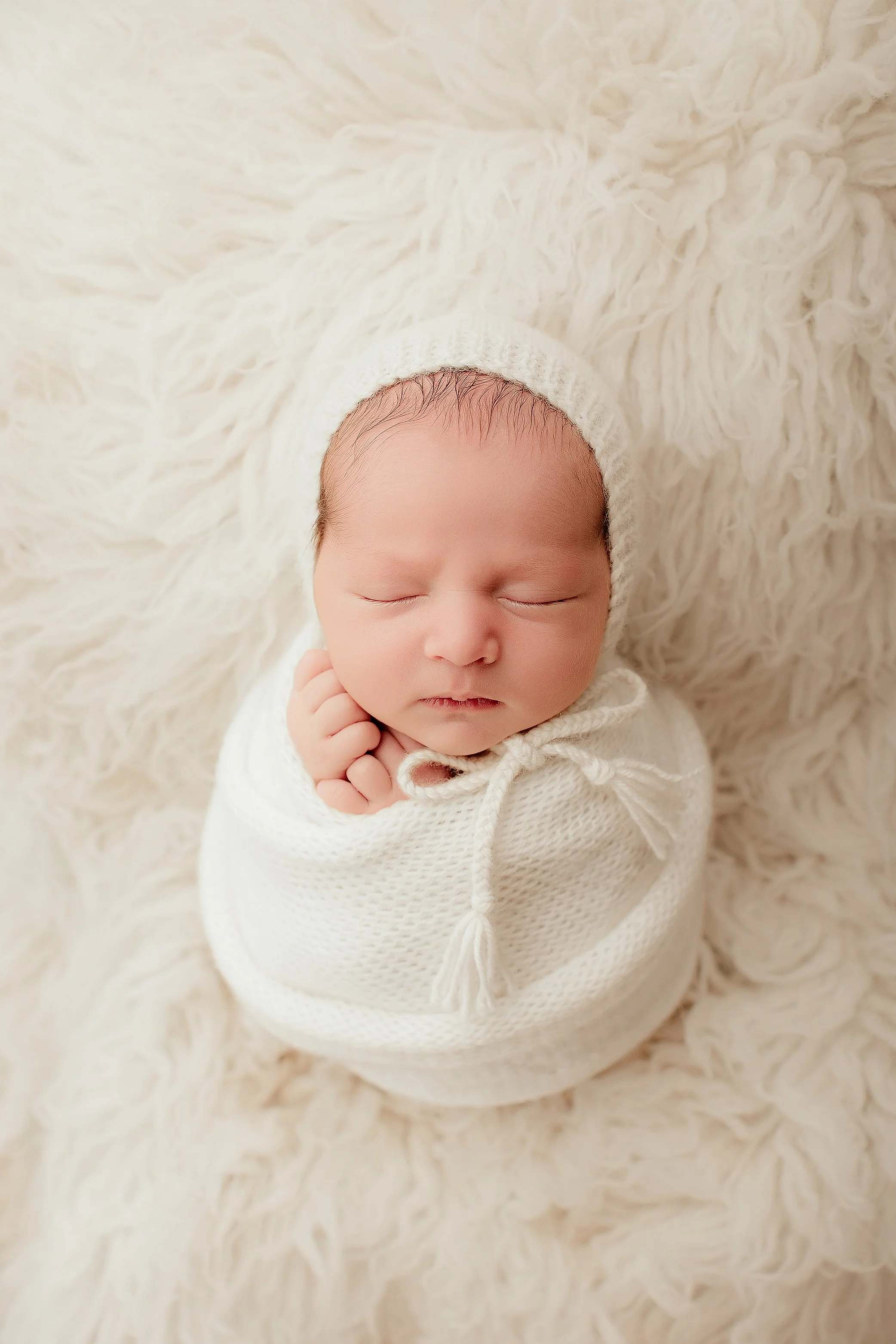 A newborn baby wrapped in a white knitted blanket and wearing a matching white knit hat, peacefully sleeping on a fluffy cream-colored surface.