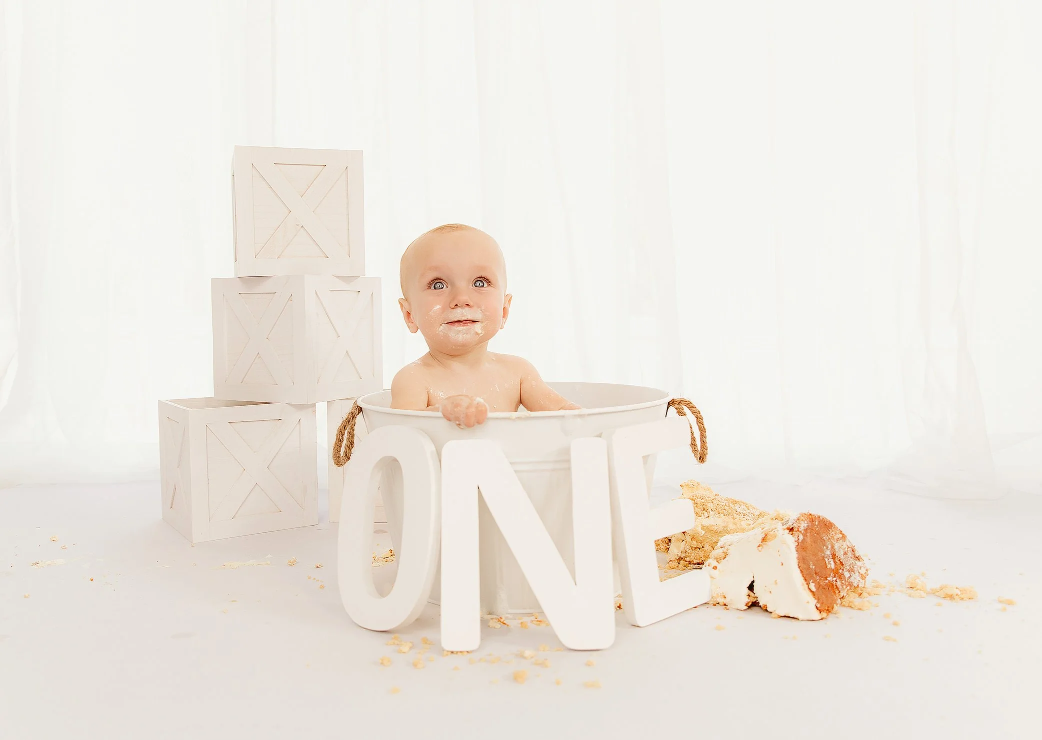 Baby in a white bucket with the word 'LOVE' on it, sitting among scattered cake crumbs and a partially eaten cake. White wooden blocks stacked behind, with sheer white curtains in background.