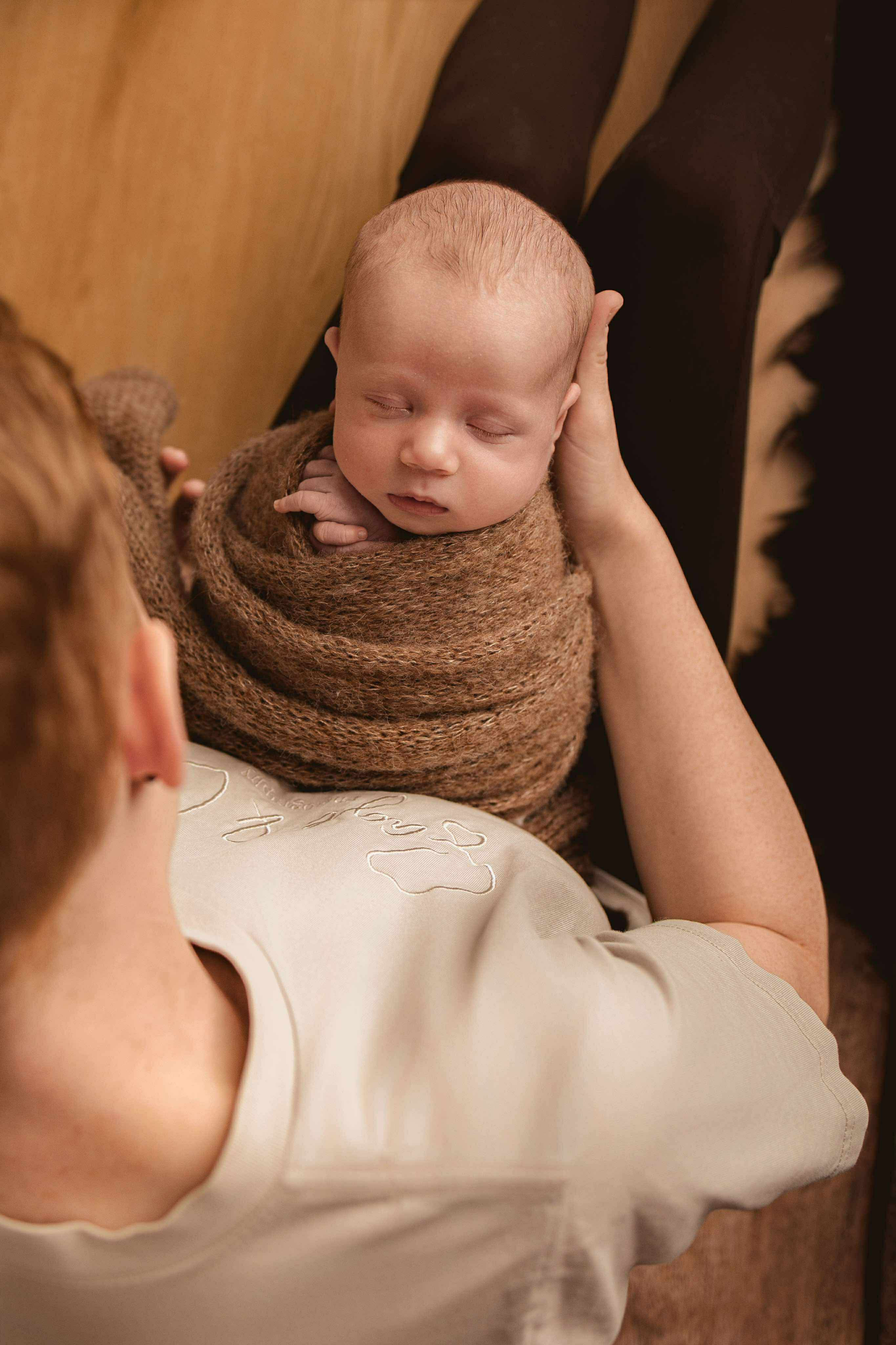 A baby wrapped in a brown blanket lying on an adult's lap, who is resting on a wooden floor. The adult is wearing a light-colored shirt with embroidery, and the scene appears cozy and intimate.