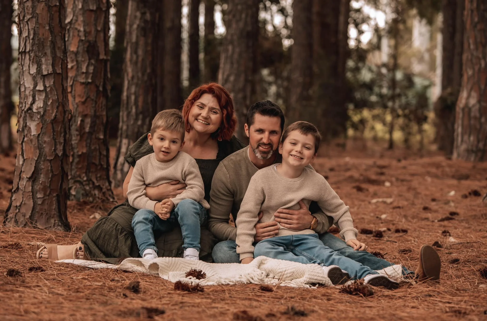 Family of four sitting on a blanket in a wooded area during fall, surrounded by tall pine trees and pine needles on the ground. They are smiling and enjoying time together.