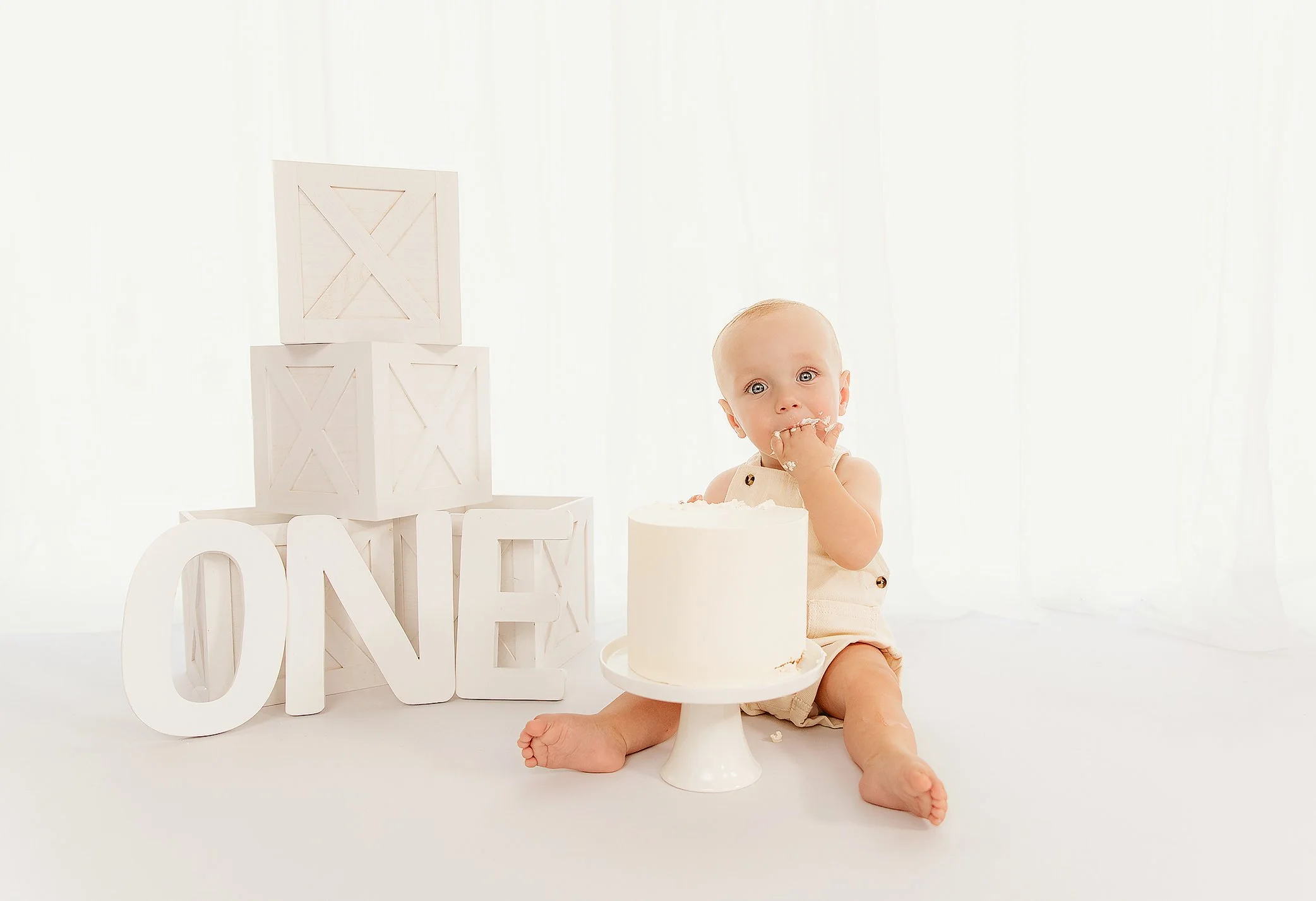 Baby sitting on the floor with a cake, surrounded by wooden blocks spelling 'ONE', celebrating a first birthday.