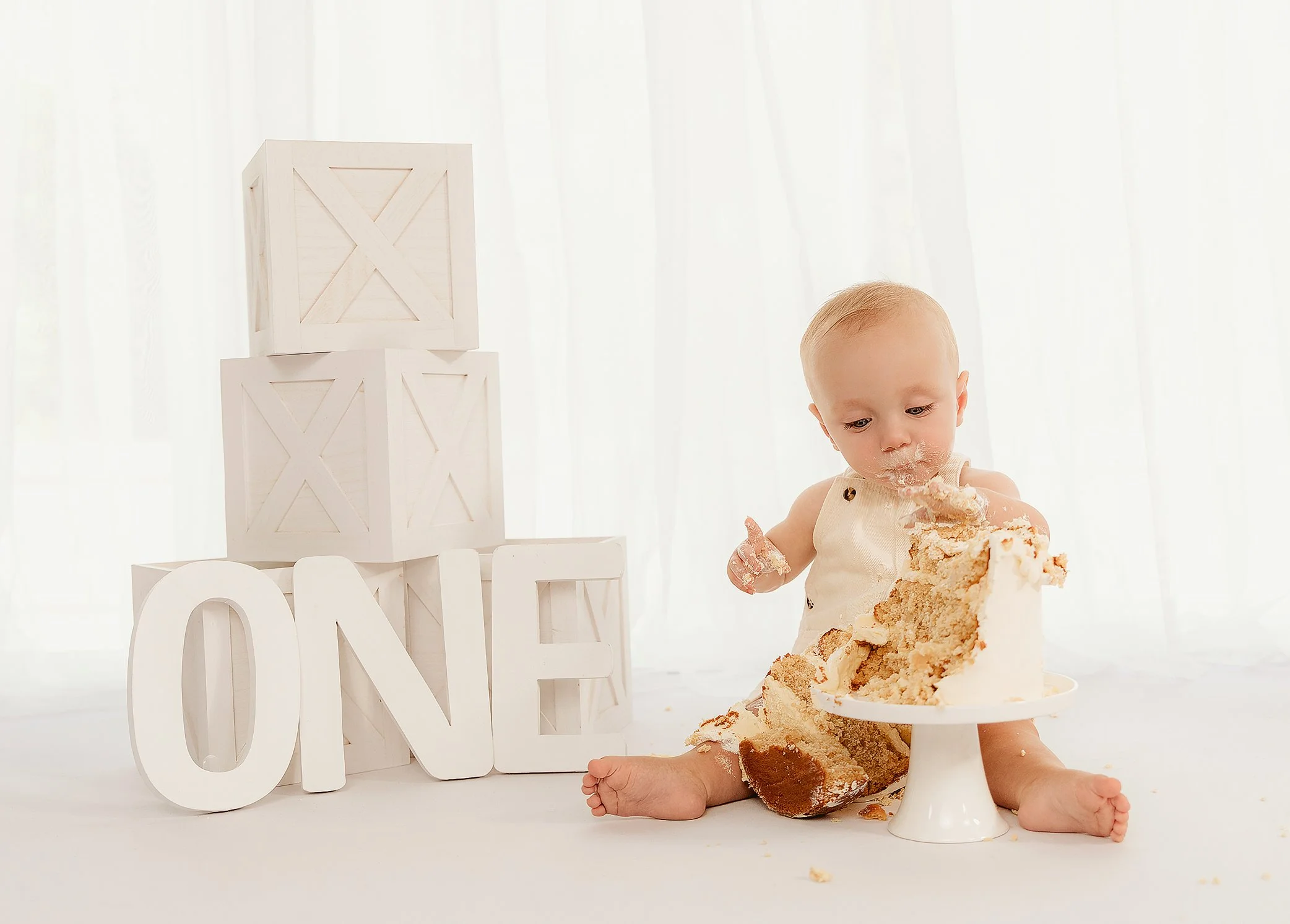 Baby sitting on a white surface, eating a large piece of cake. There are white decorative blocks spelling 'ONE' and some white crates in the background.
