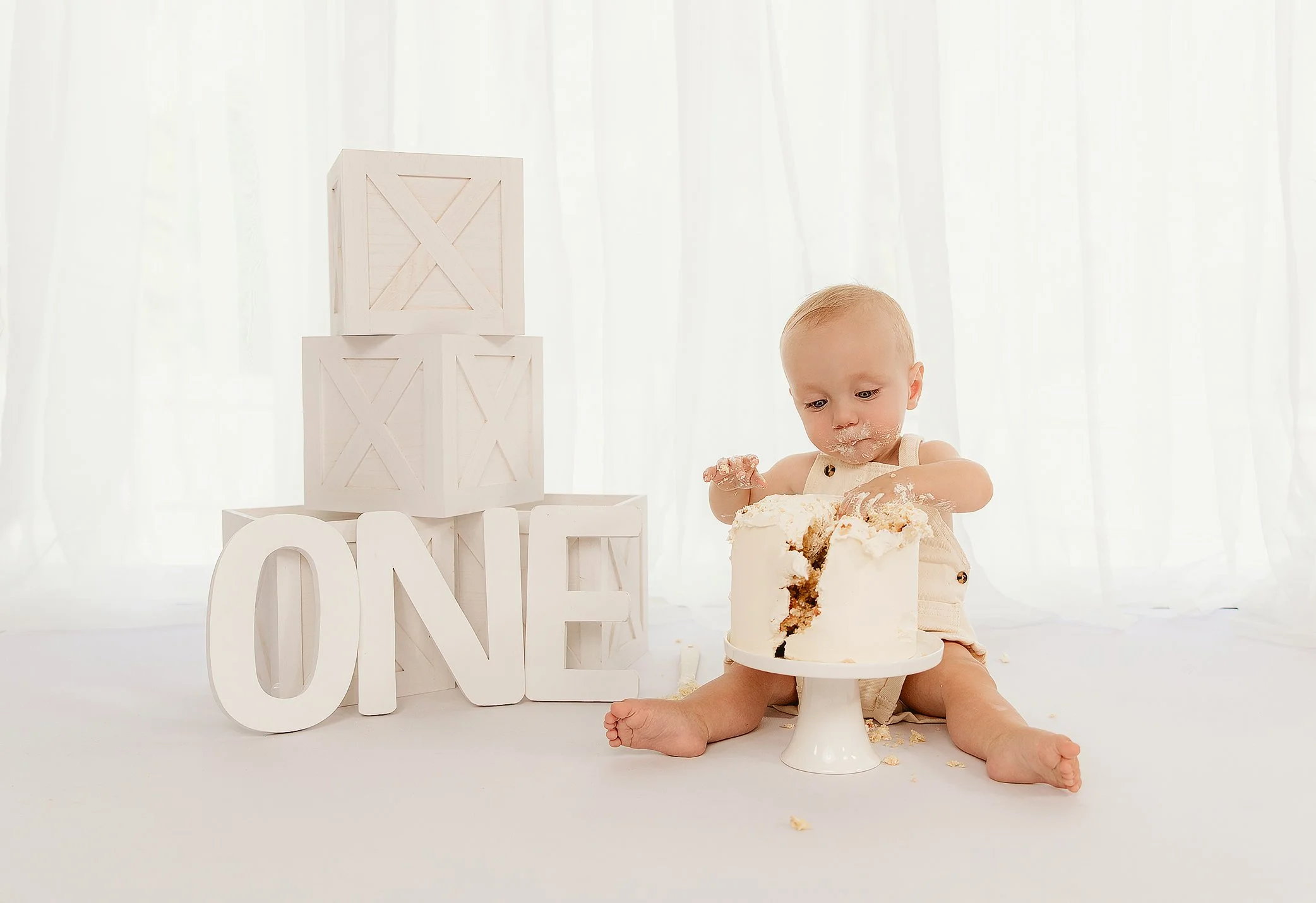 Young child sitting on the floor with a partially eaten cake on a white cake stand during a celebration, with decorative blocks spelling 'ONE' beside them, in a bright room with white curtains.