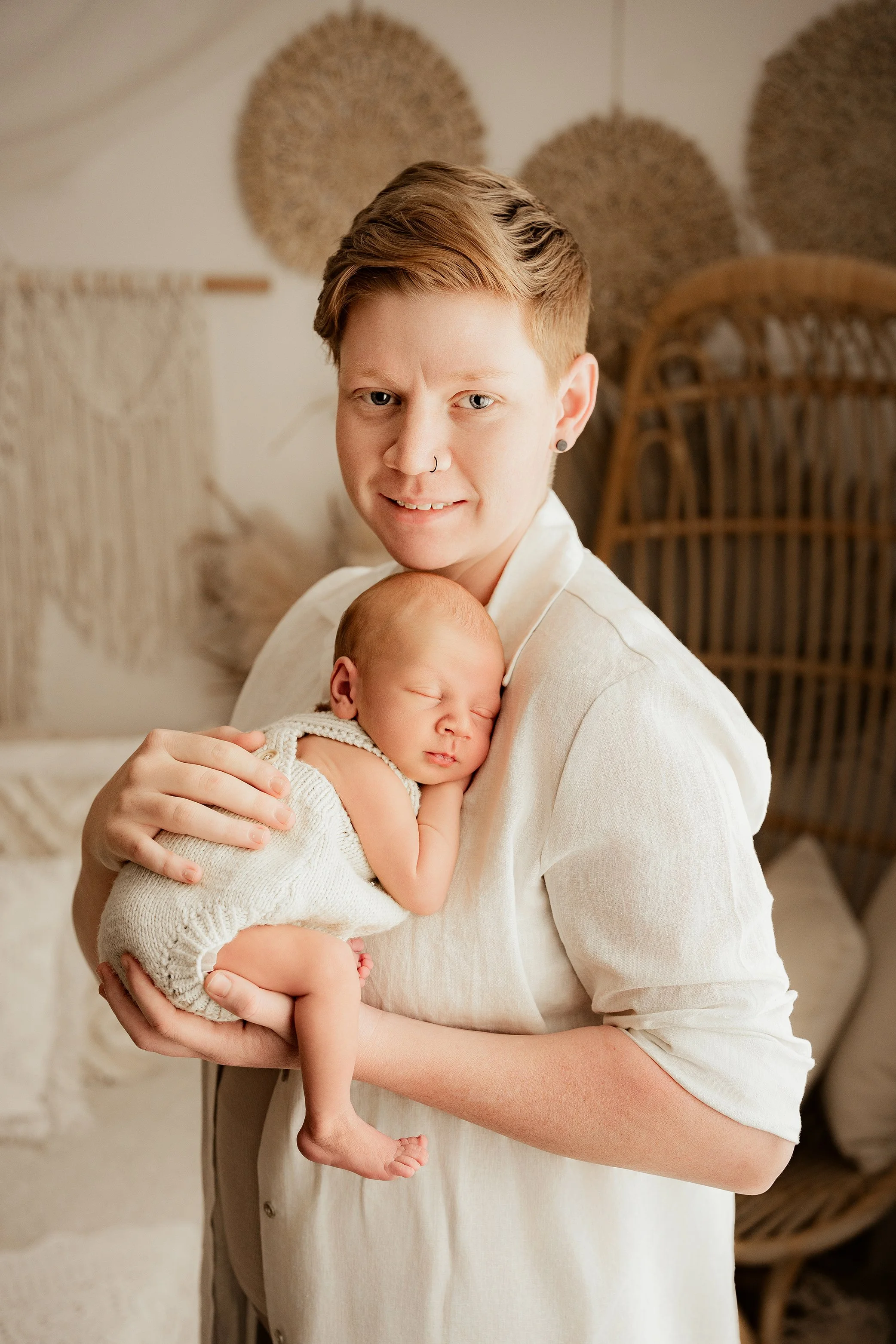 A person with short hair and a nose ring holds a sleeping baby dressed in a knit white romper in a warmly decorated room.