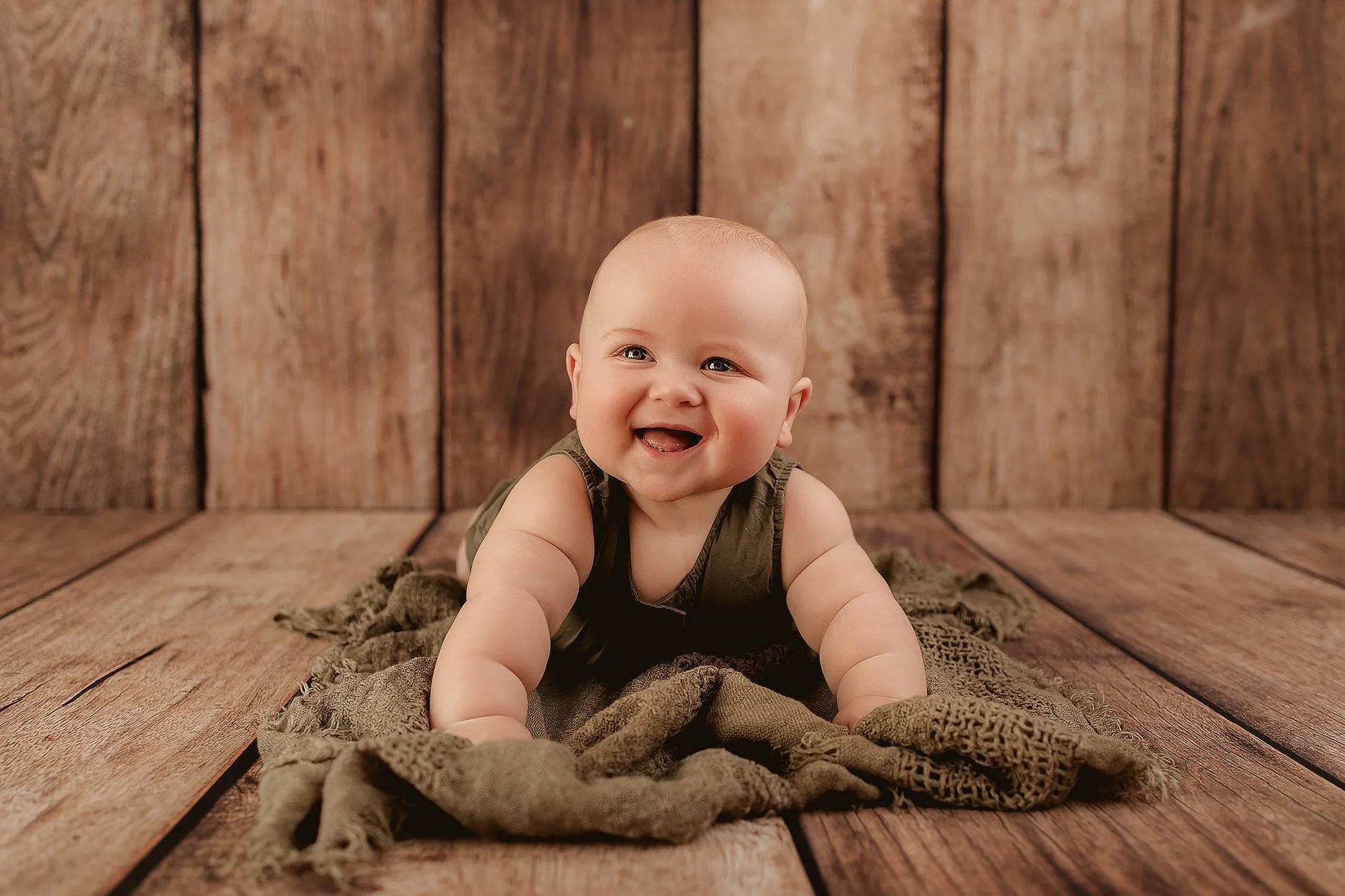 Smiling baby crawling on a wooden floor with a textured blanket, wooden wall background.