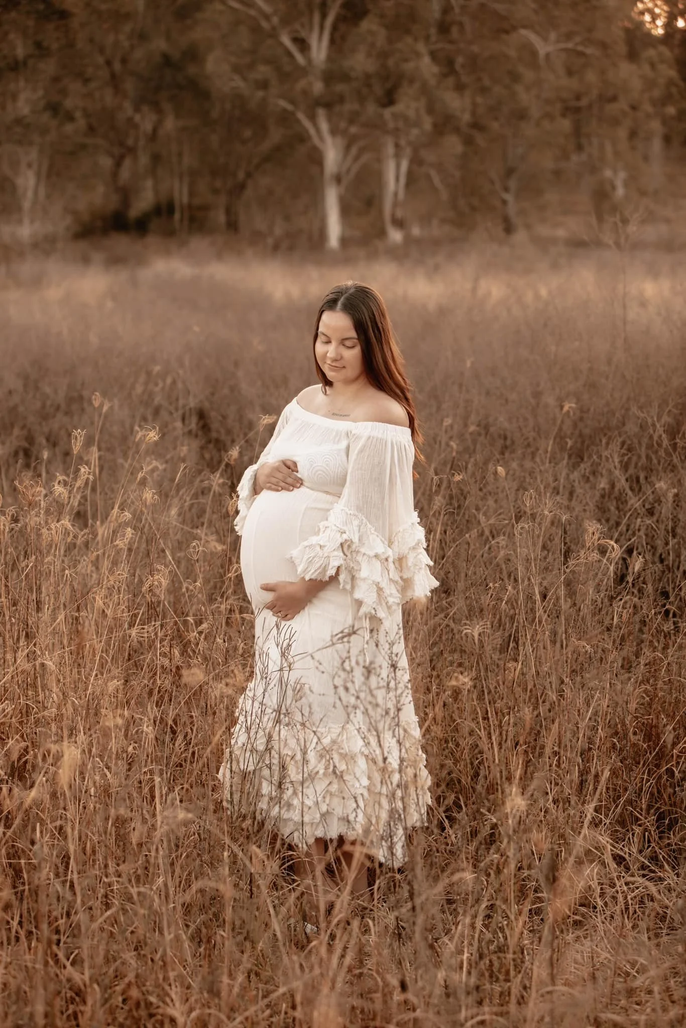A pregnant woman in a white dress with ruffled sleeves standing in a golden field of tall grass during sunset, gently holding her belly with one hand and her other hand on her shoulder, with trees in the background.