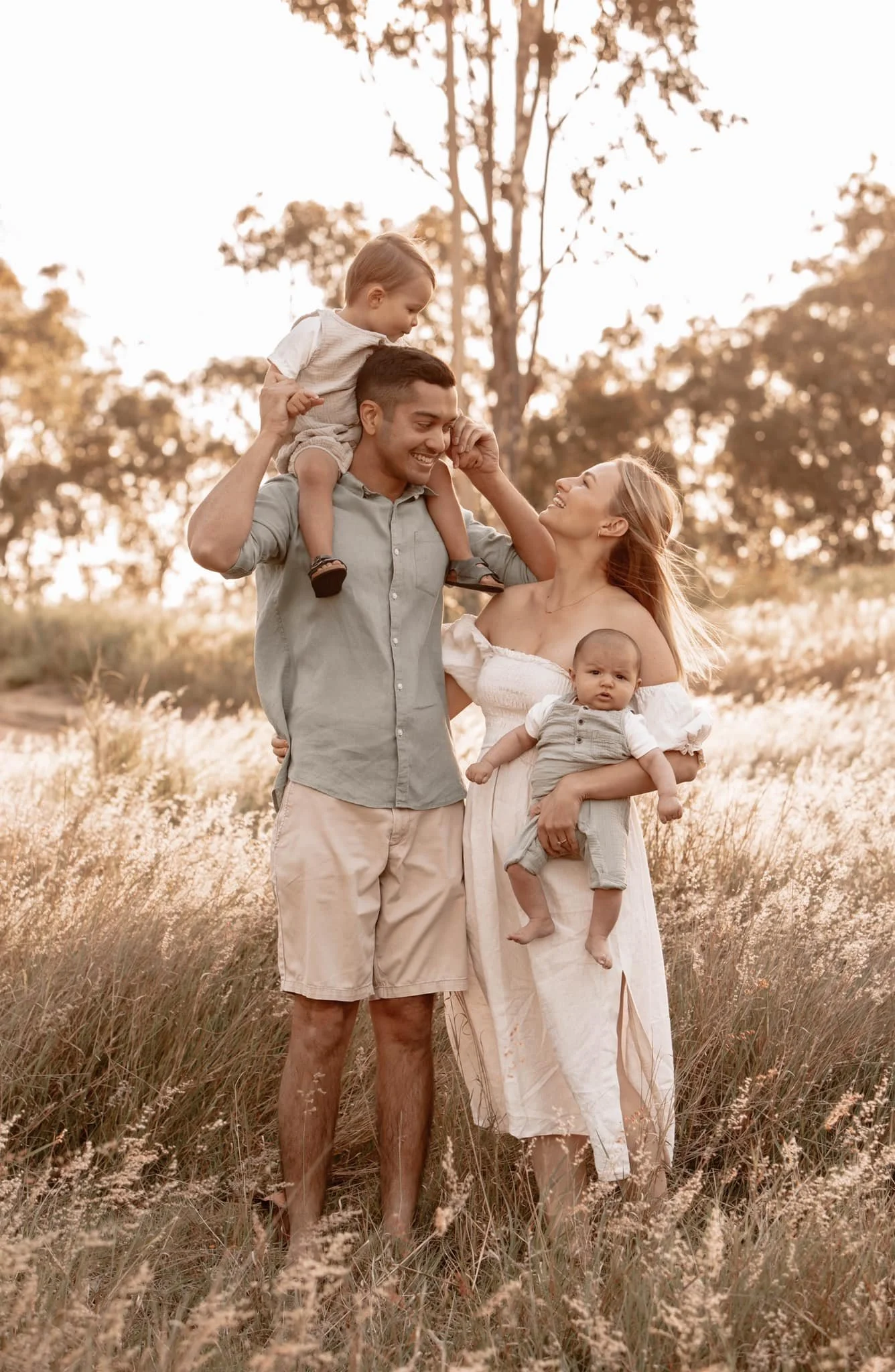 A happy family of four outdoors in a field during sunset, with the father carrying a toddler on his shoulders, the mother holding a baby, all smiling and looking at each other.