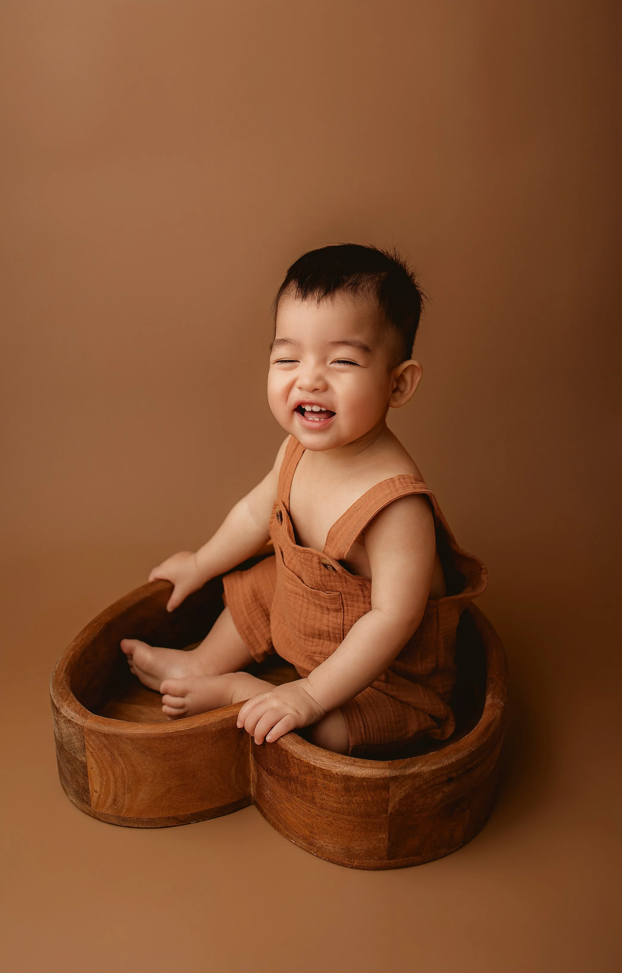 Smiling young boy sitting in a wooden container against a brown background, wearing brown overalls.