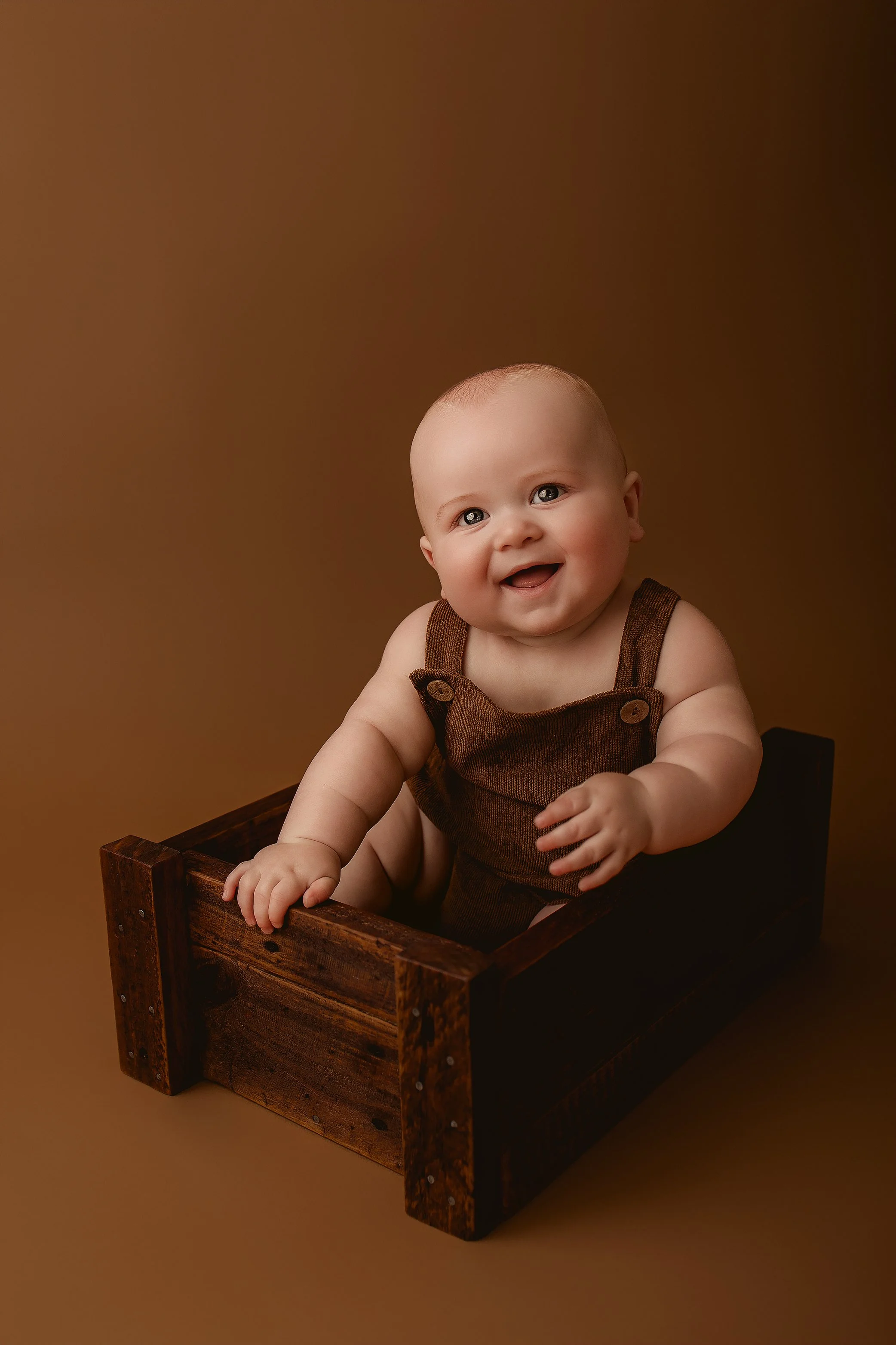 A happy baby sitting inside a wooden box, wearing brown overalls, against a brown background.