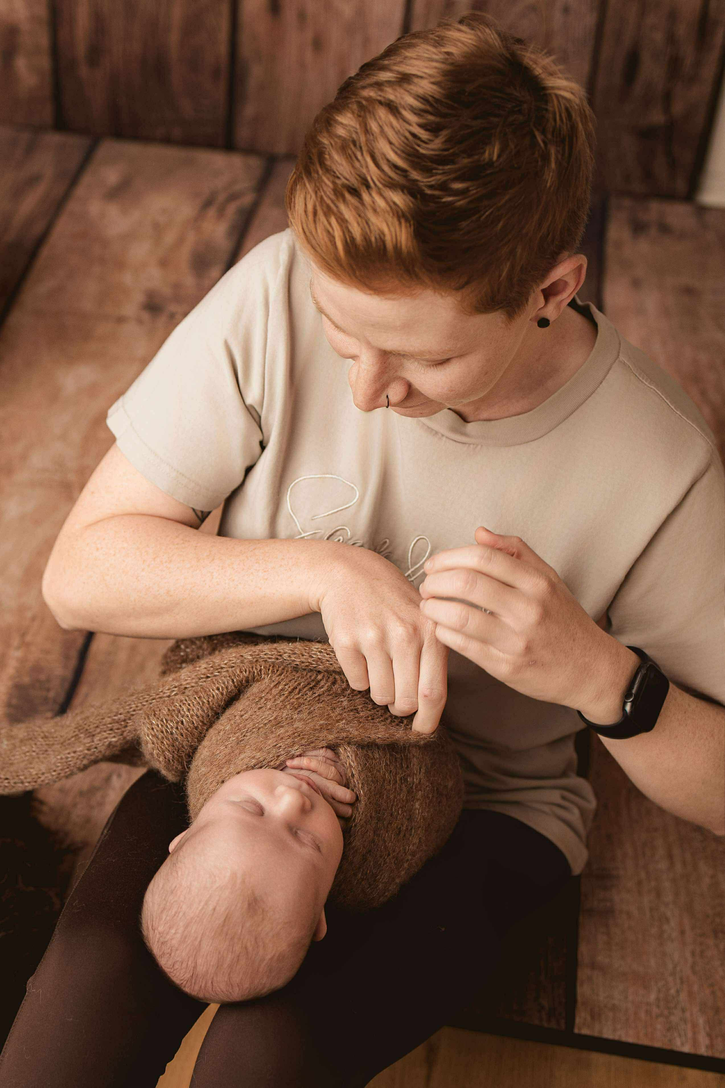 A person with short red hair and wearing a beige T-shirt is lying on a brown bench while cradling a sleeping infant wrapped in a brown knitted blanket on their chest.