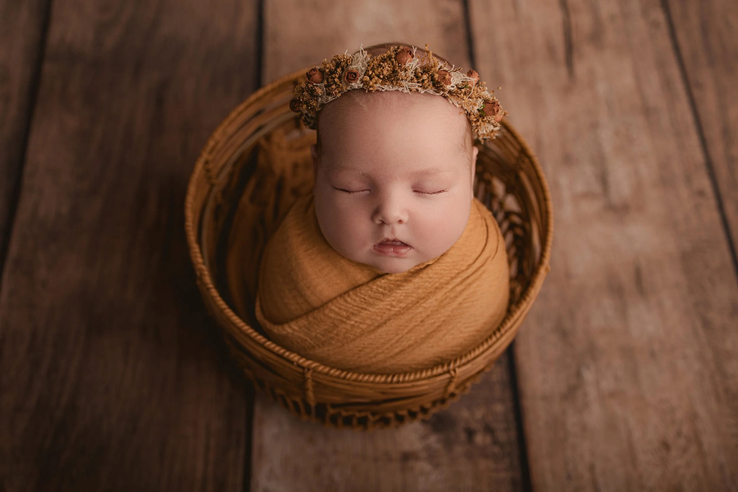 A sleeping baby in a woven basket, wrapped in a caramel-colored blanket, wearing a floral headband, on a wooden floor.