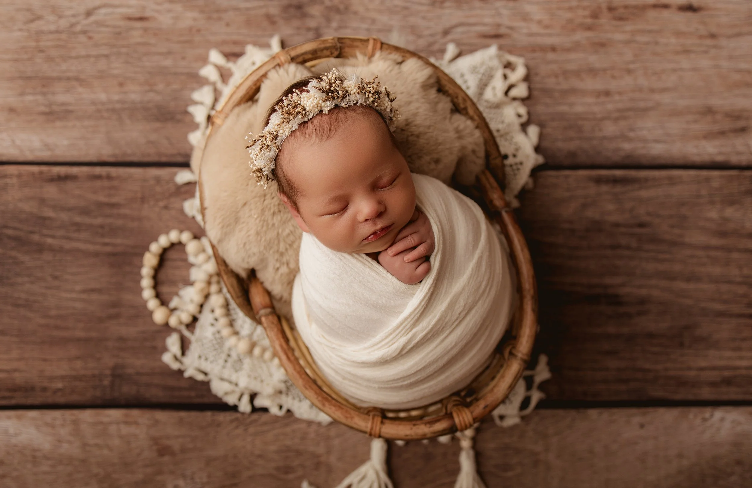 A sleeping newborn baby swaddled in white wraps inside a wicker basket with a lace cloth underneath, wearing a floral headband, on a wooden floor.