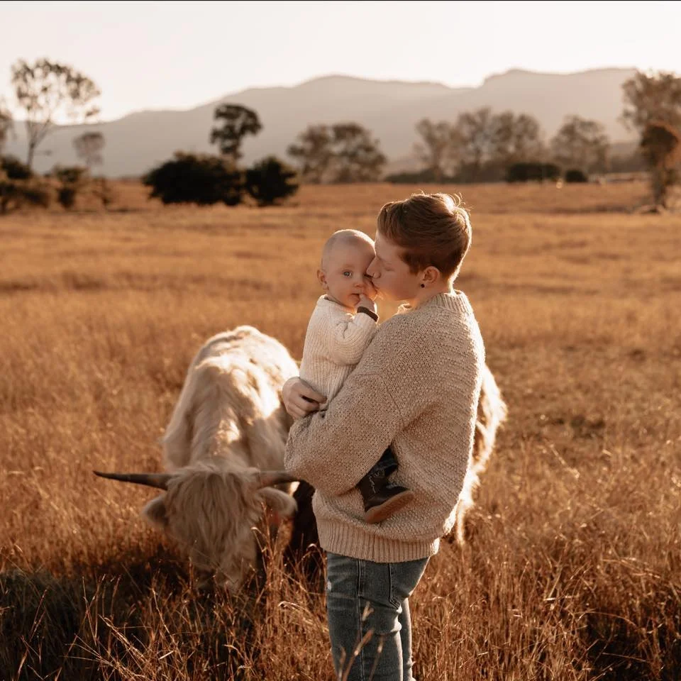 A woman holding a young child in a field with a grazing Highland cow. The scene is during sunset with mountains and trees in the background.