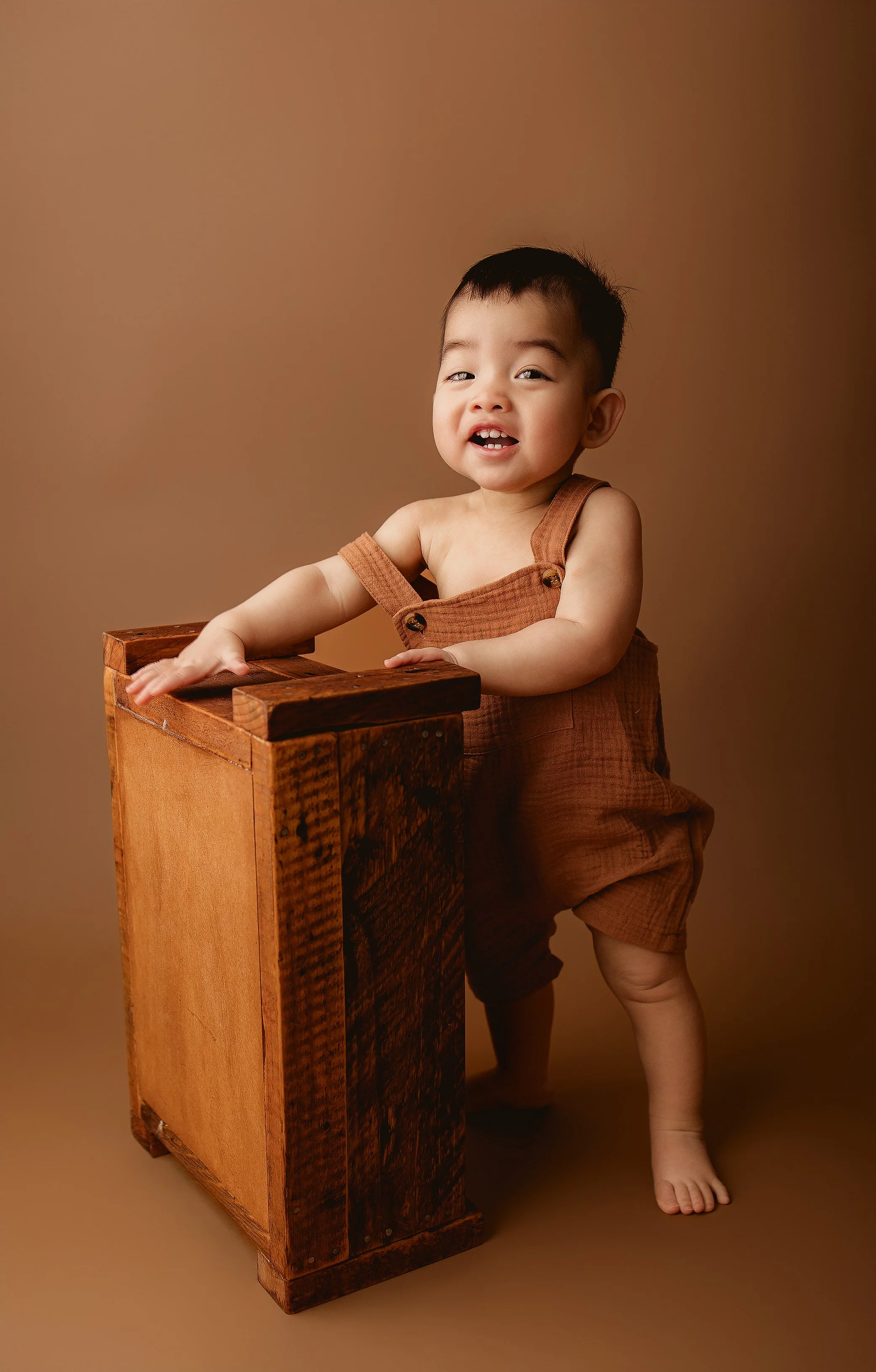 Young child with short dark hair, wearing brown overalls, standing barefoot and leaning on a wooden block against a plain brown background.