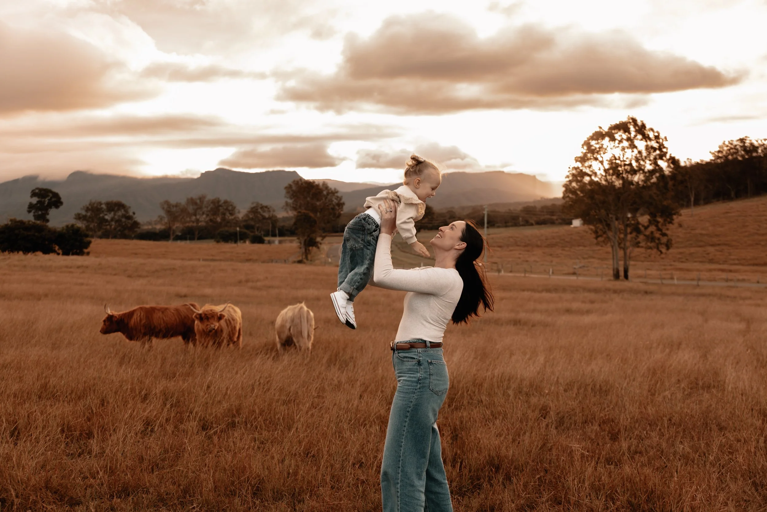 A woman lifts a young girl in the air while standing in a grassy field with cows grazing, trees, mountains, and a cloudy sky in the background during sunset.