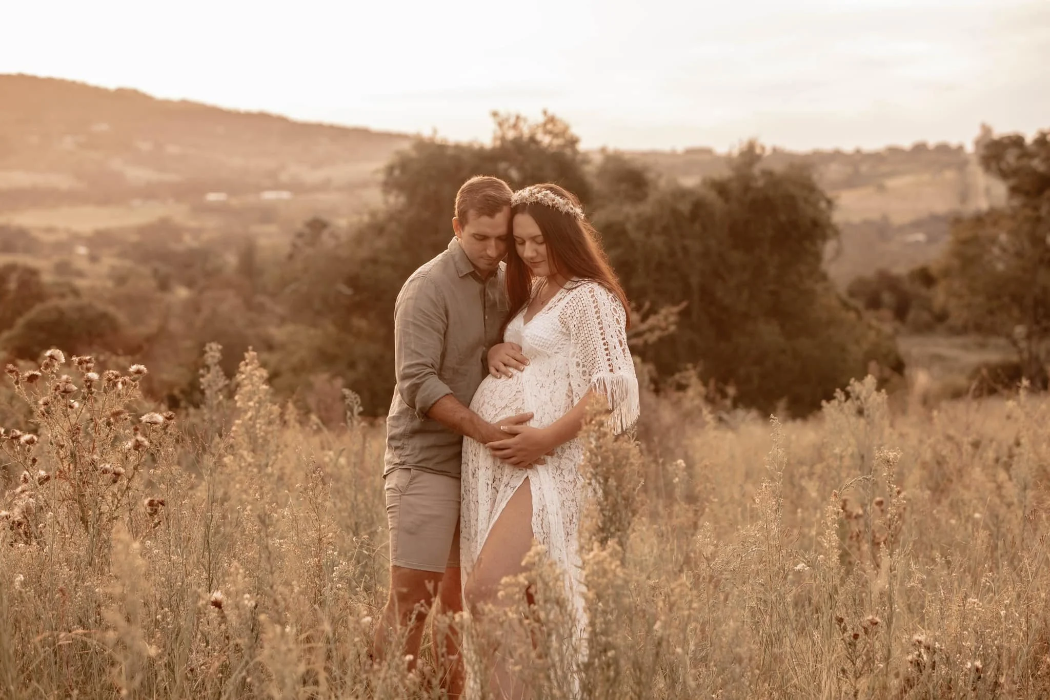 Couple standing in a field during sunset, the man gently holding the pregnant woman's belly as they look down, surrounded by tall grass and wildflowers.