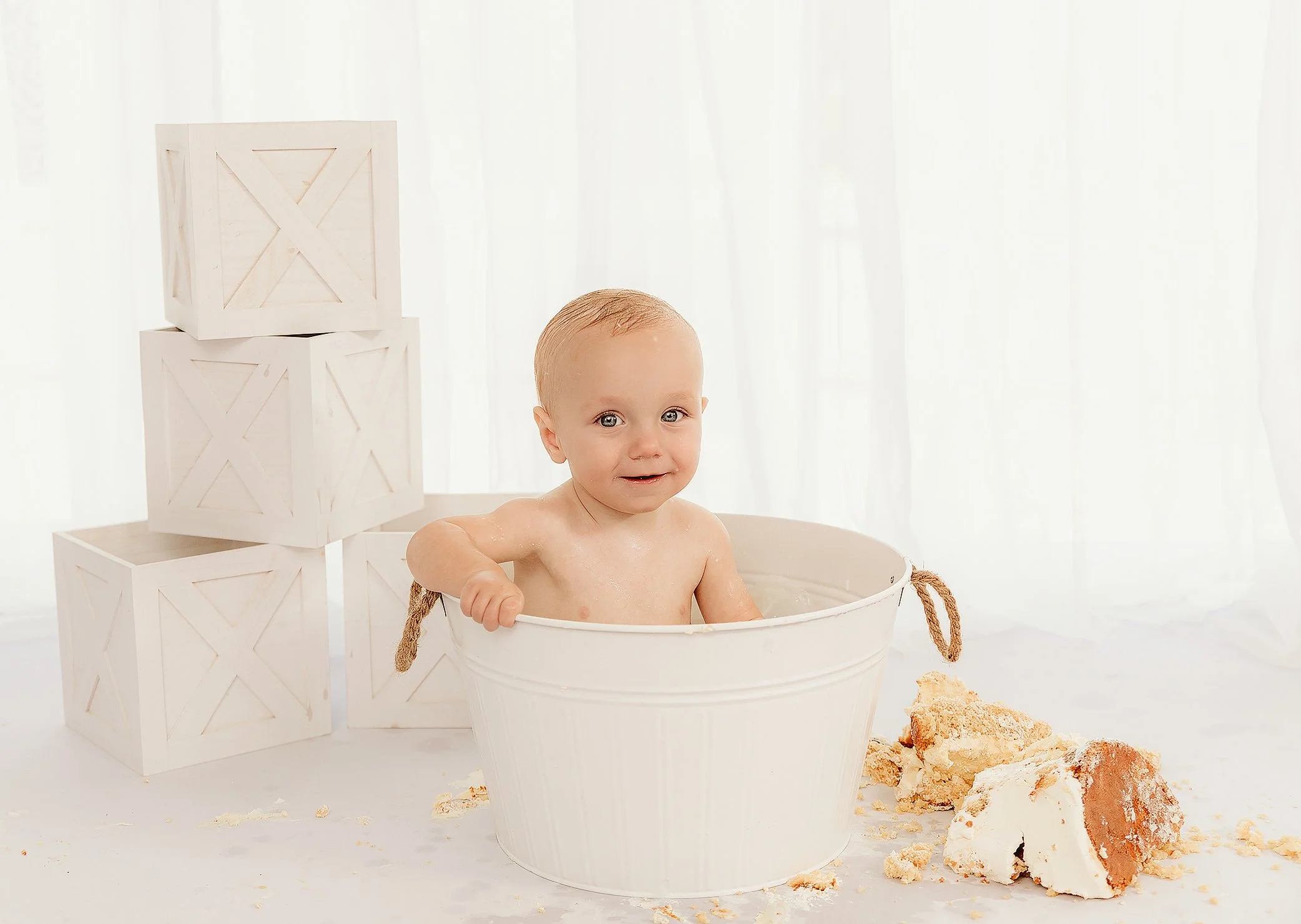 A young child with wet hair sitting in a white tub of water, surrounded by cake crumbs, with white crates stacked nearby and sheer white curtains in the background.