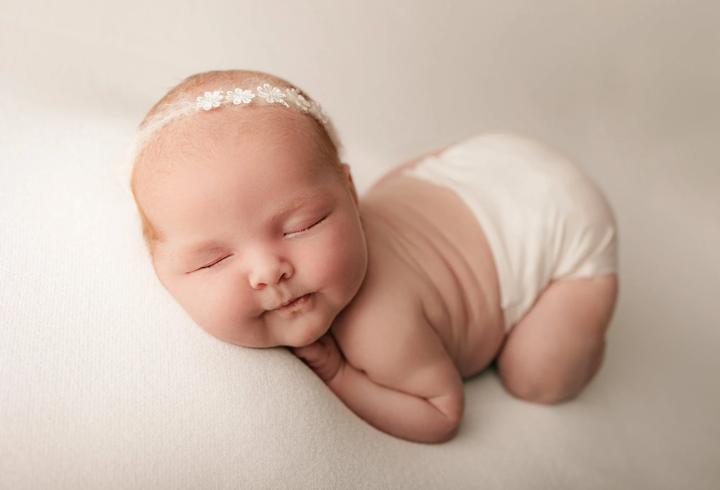 A sleeping baby with a floral headband, lying on its side on a soft, light-colored surface.