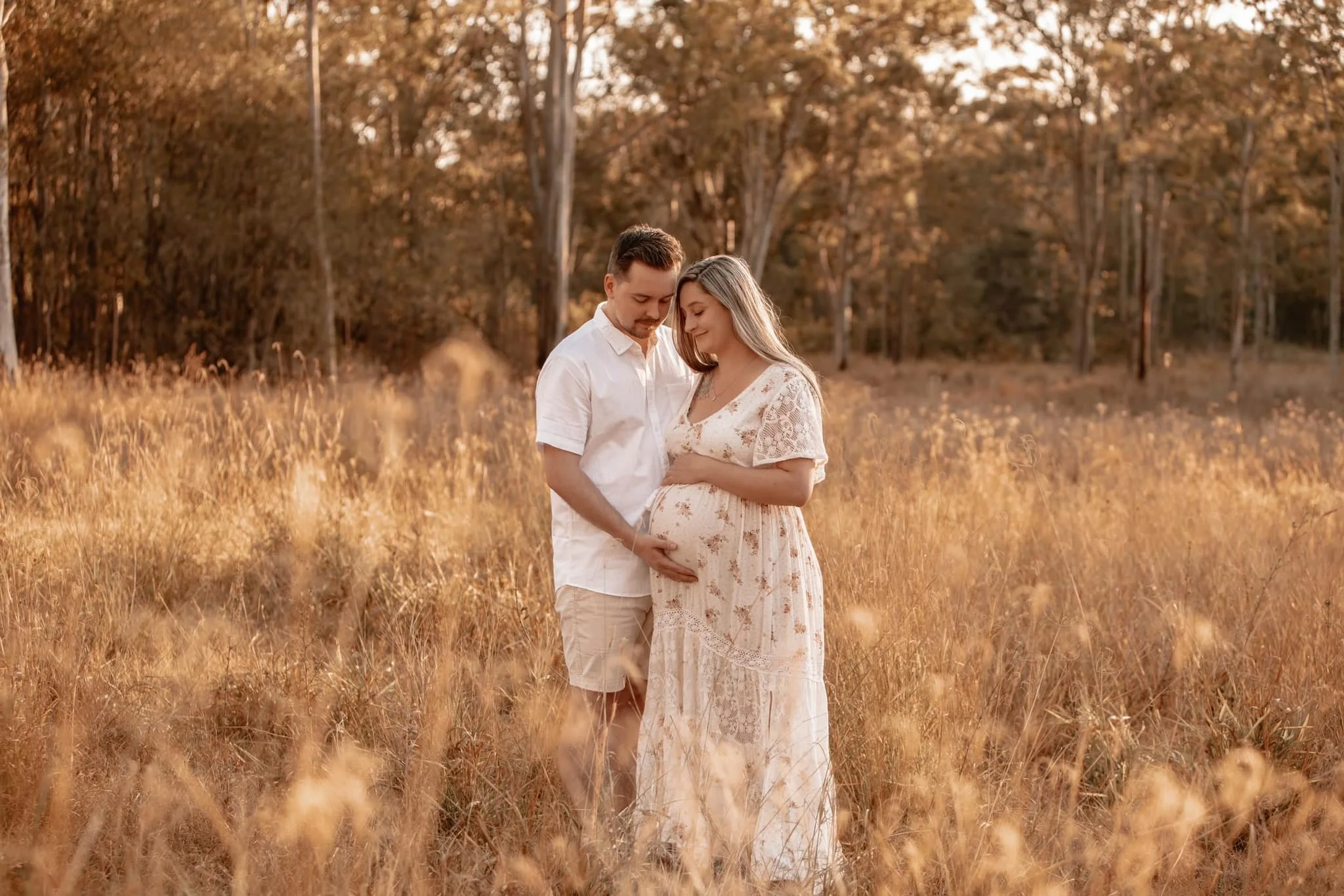 A pregnant woman and a man standing in a field of tall grass during sunset, looking at her baby bump with affection.