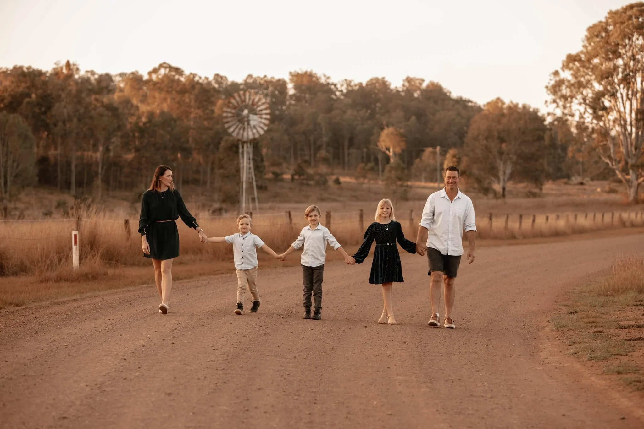Family of six walking hand in hand on a dirt road during sunset, with trees and a windmill in the background.