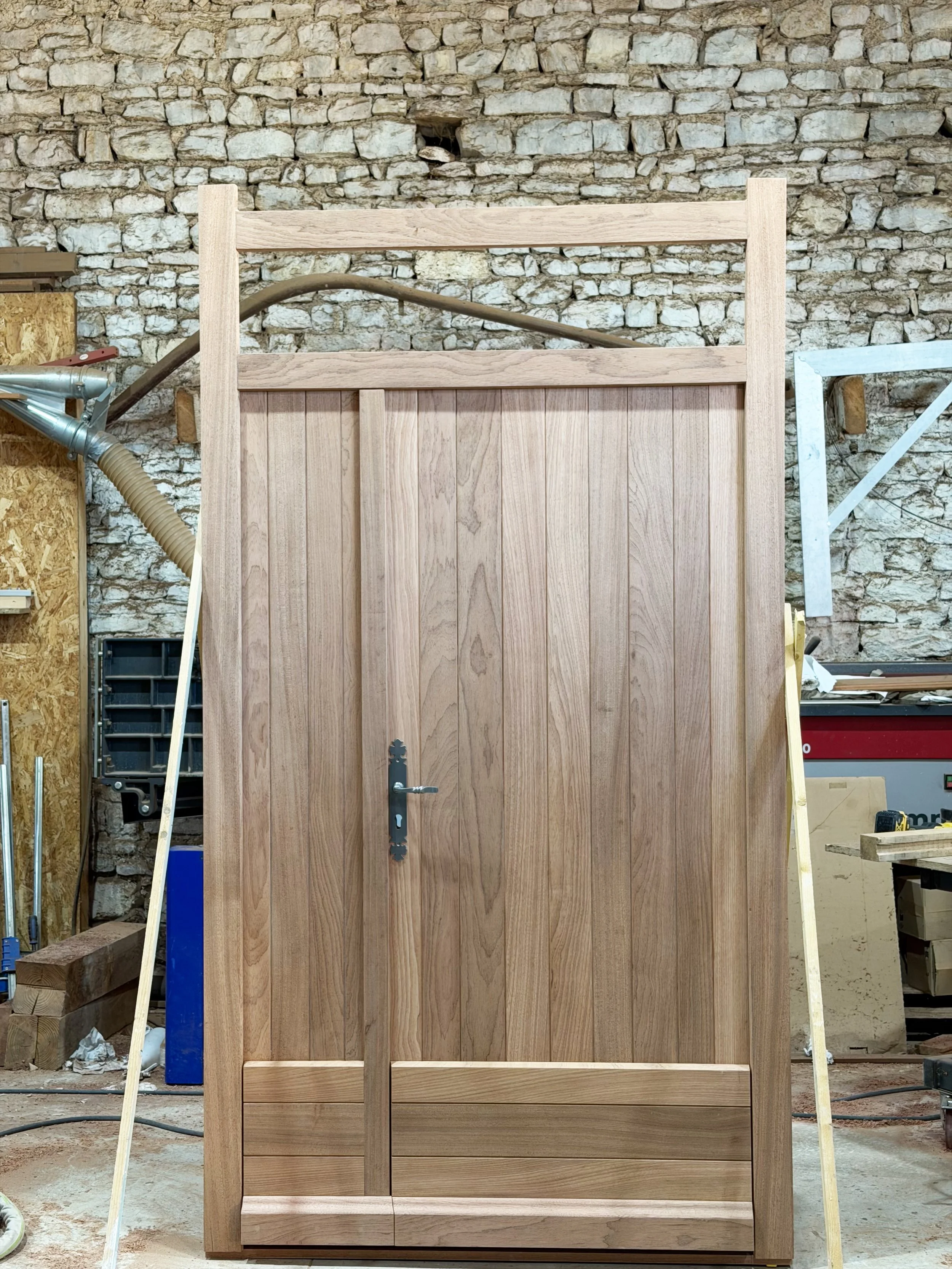 Portes doubles pour une maison à Dirac (16), photographiées à l'atelier avant vitrage et mise en peinture. Vantaux à lames verticales, imposte vitrée et dormant fabriqués d'un seul tenant, poignée en fer forgé.
