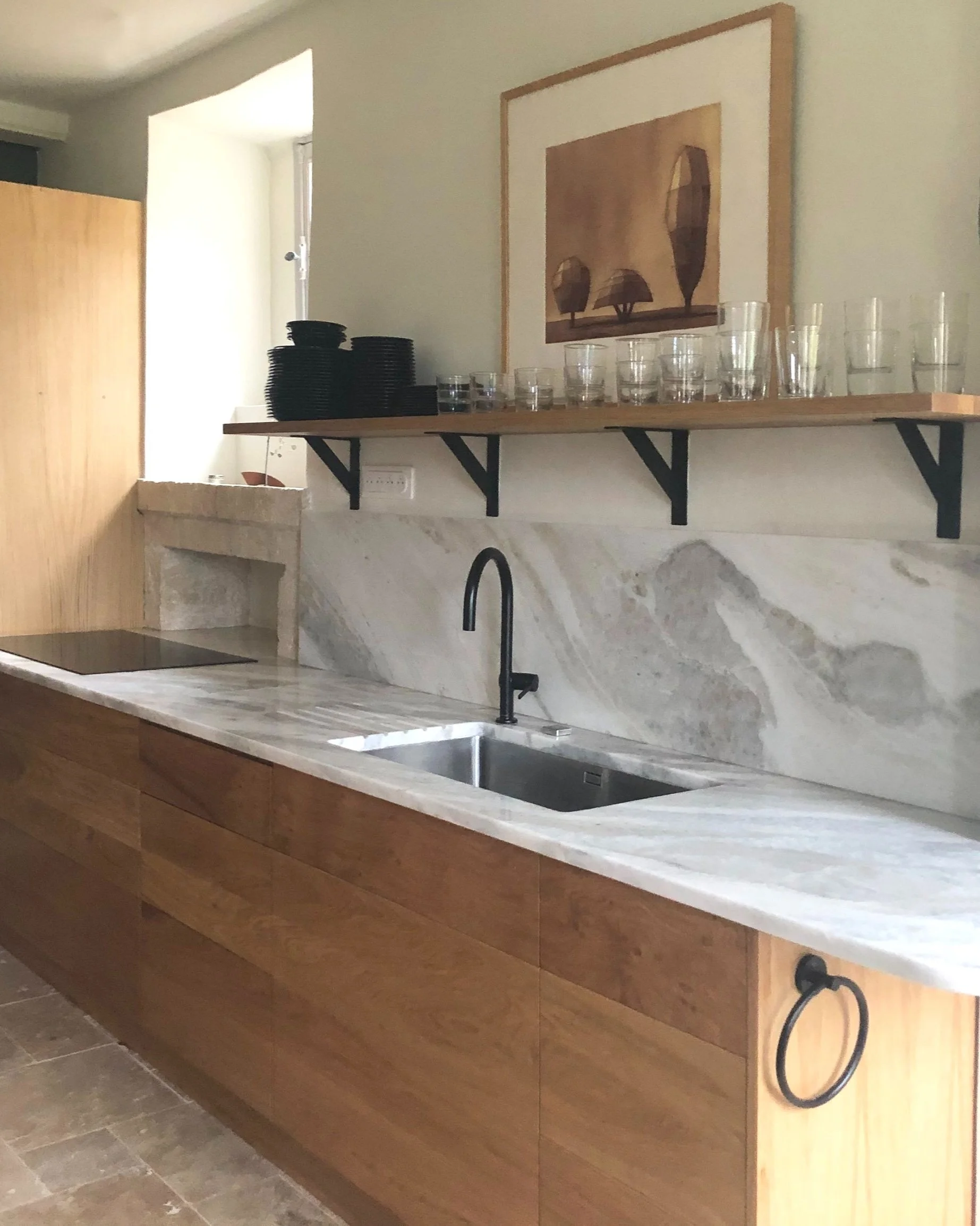 Cuisine avec plan de travail en marbre, évier avec robinet noir, étagère en bois avec verres et assiettes, peinture d'arbres sur le mur.