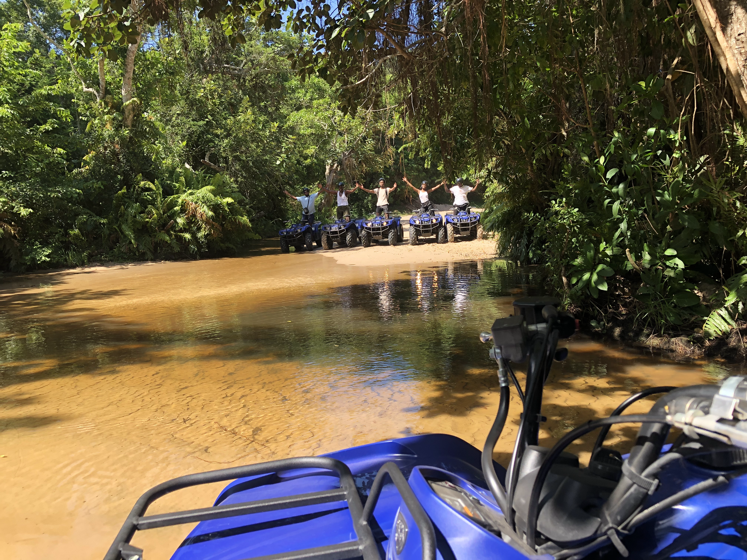 A group of people riding ATVs through a jungle river, with lush green trees and vegetation surrounding them.