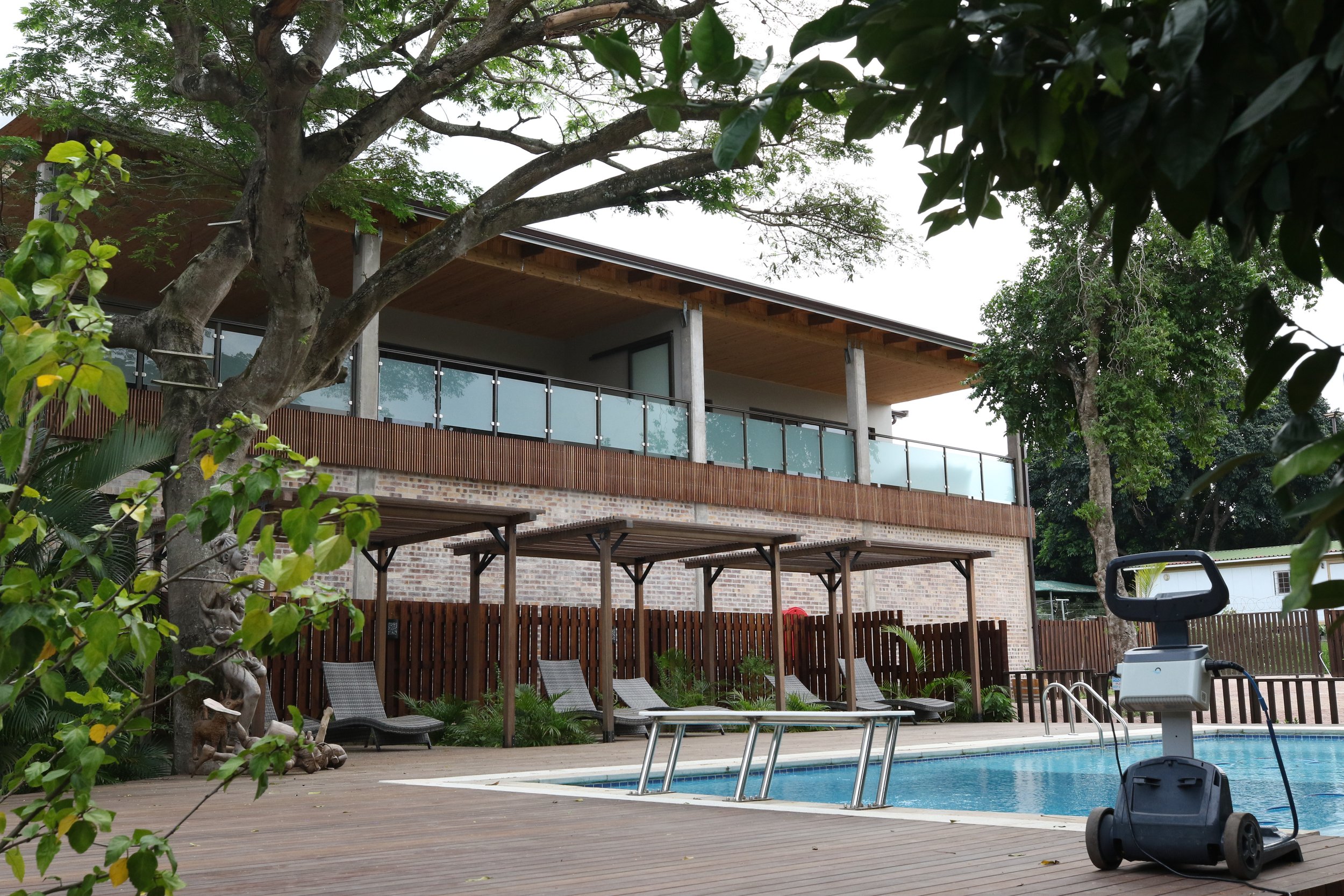 A backyard scene featuring a swimming pool with a pool cleaning robot, pool skimmer, and two lounge chairs under a small shaded area. Tall trees surround the yard, with a modern house with large windows and balcony in the background. The scene is enclosed by a wooden fence.