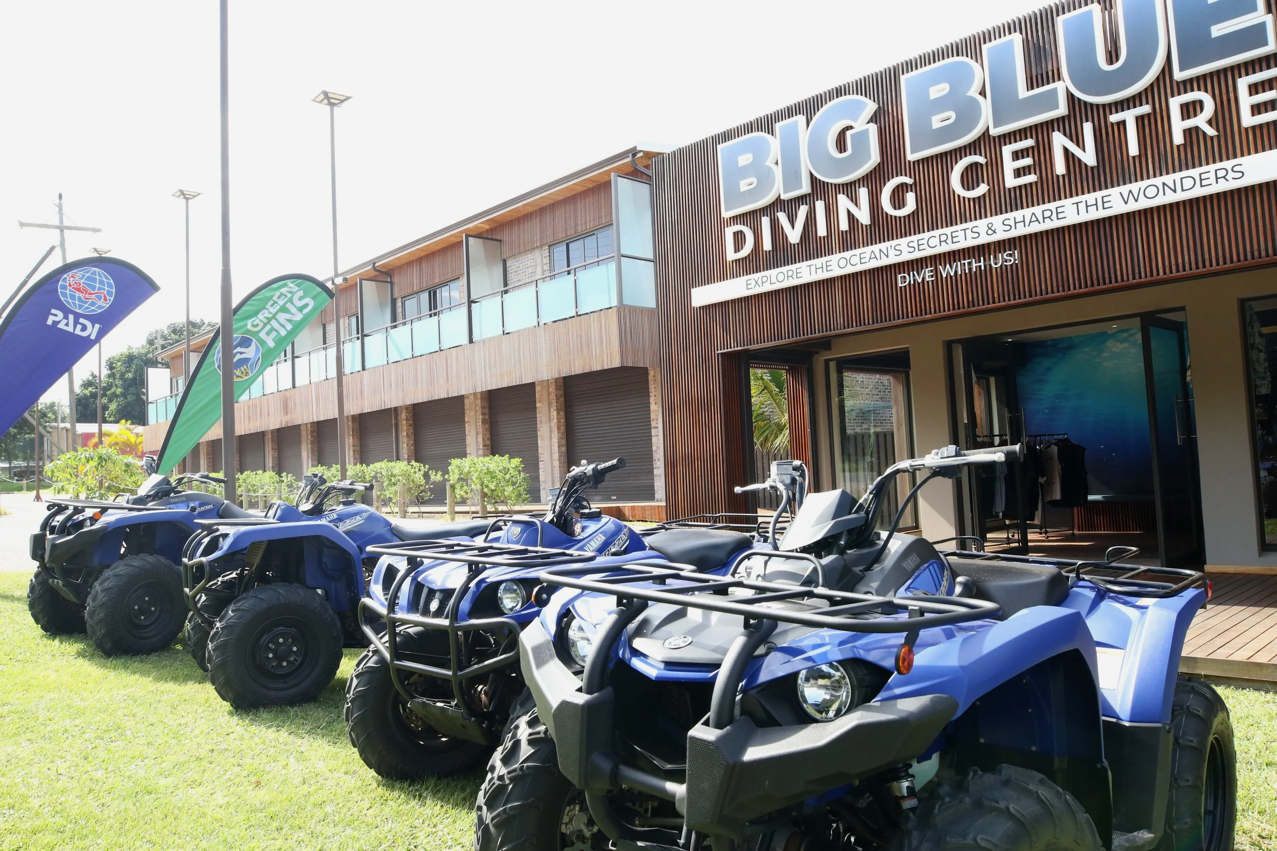 A row of blue and white all-terrain vehicles parked on a grassy area in front of a building with a sign that reads Big Blue Diving Centre, with flags that display PADI and Green Fins logos.