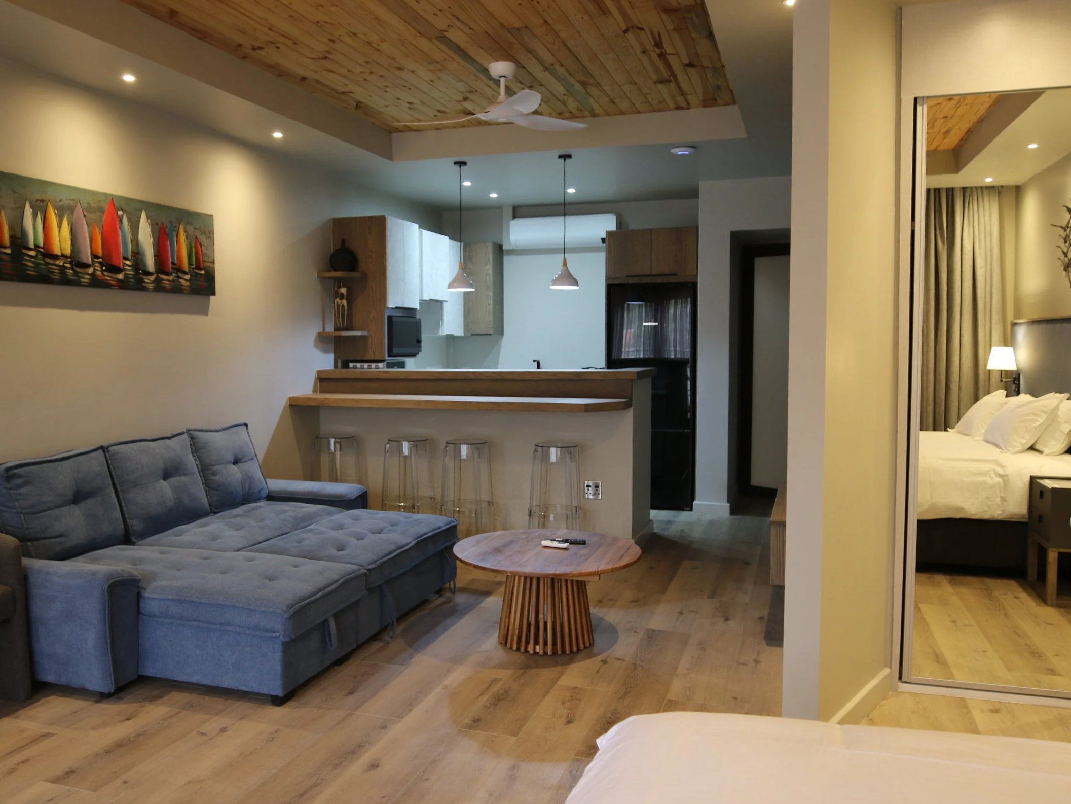 Open-concept living area with a gray sofa, wooden coffee table, kitchen with pendant lights, bar stools, and a colorful boat-themed wall art.