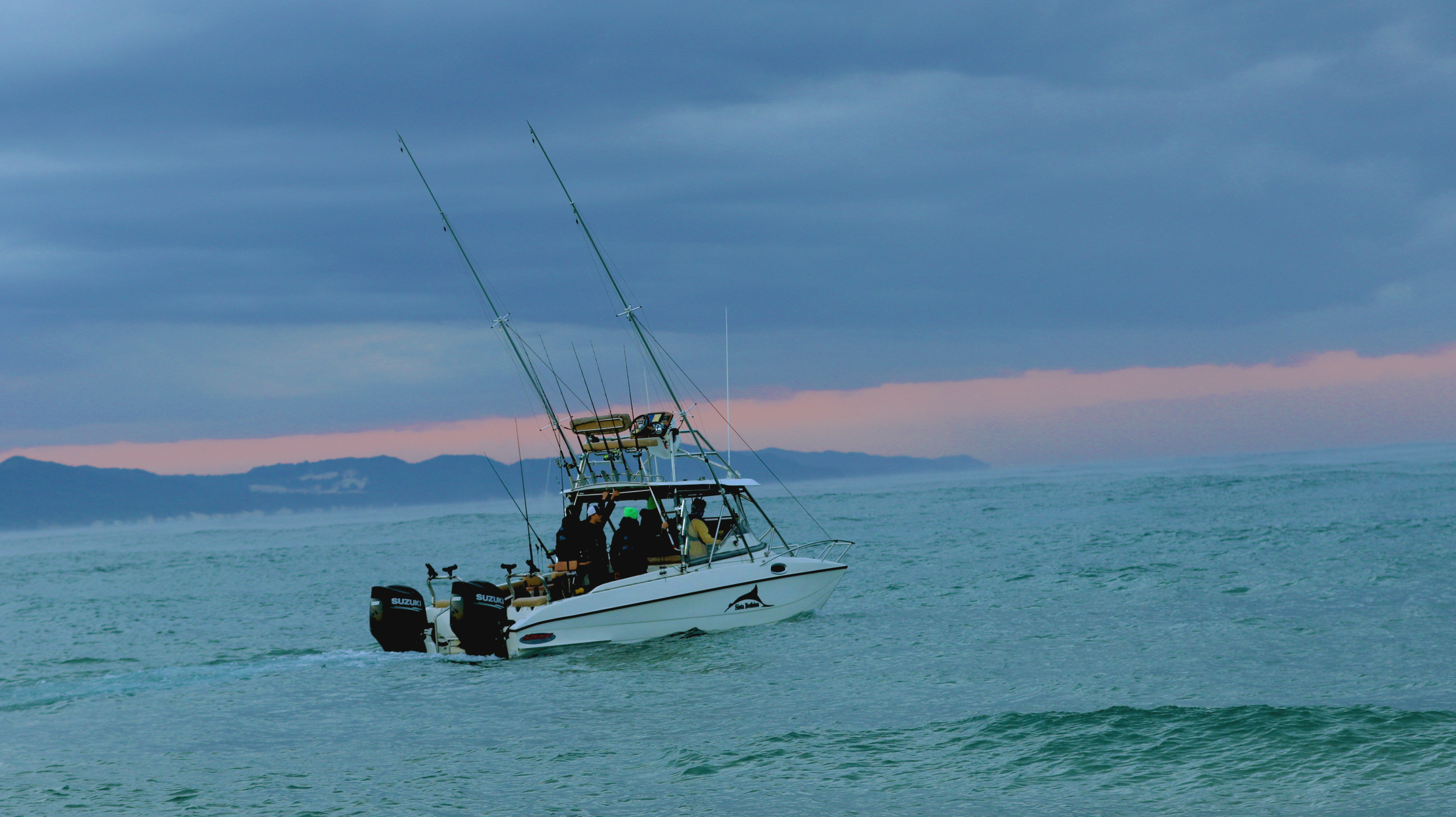 A fishing boat sailing on calm ocean waters under a cloudy sky at dusk, with mountains visible in the distance.
