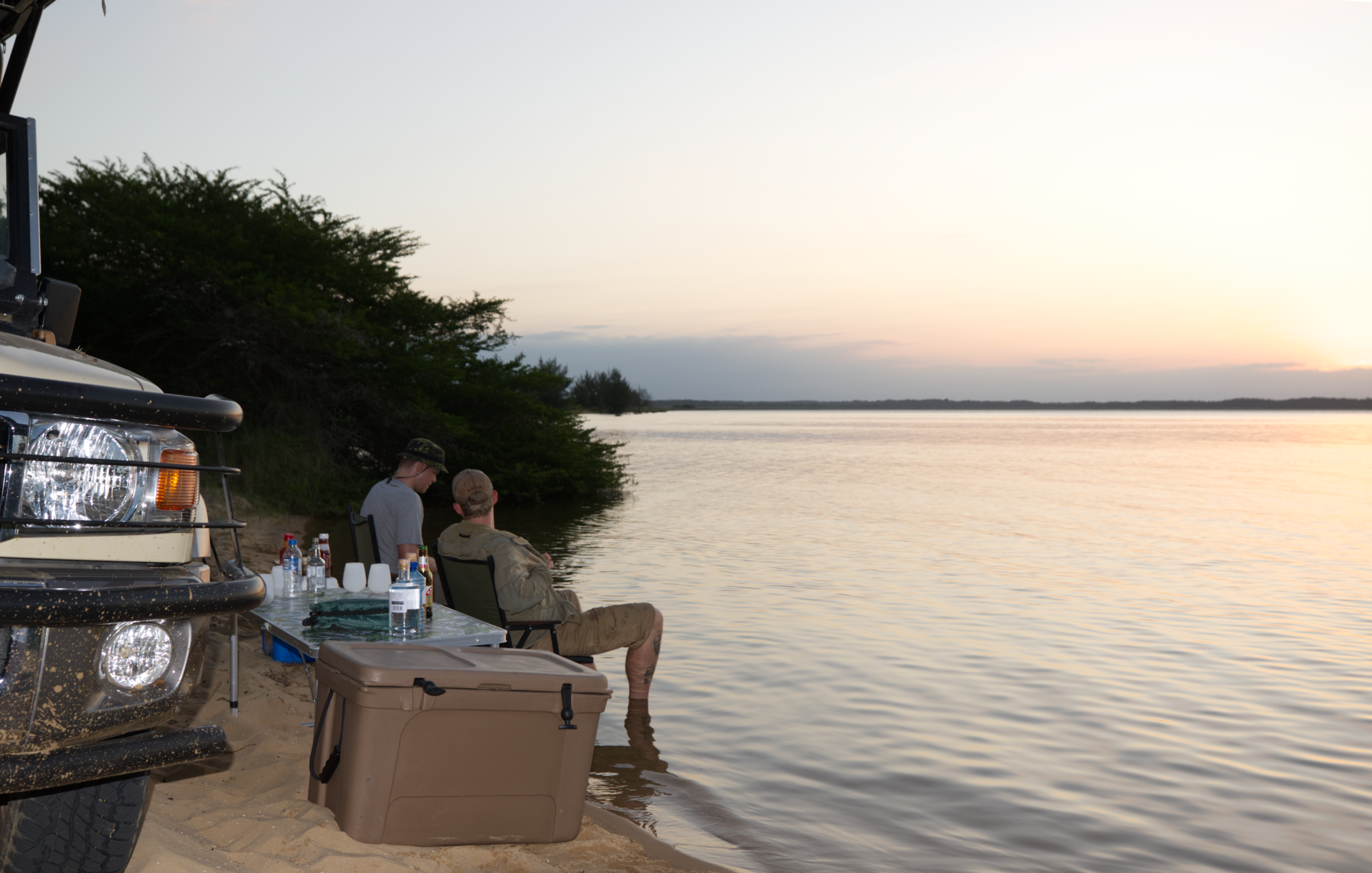 Two men sitting by the shore of a large body of water at sunset, with a vehicle and a table with bottles and cups nearby, and a large cooler in front of them.