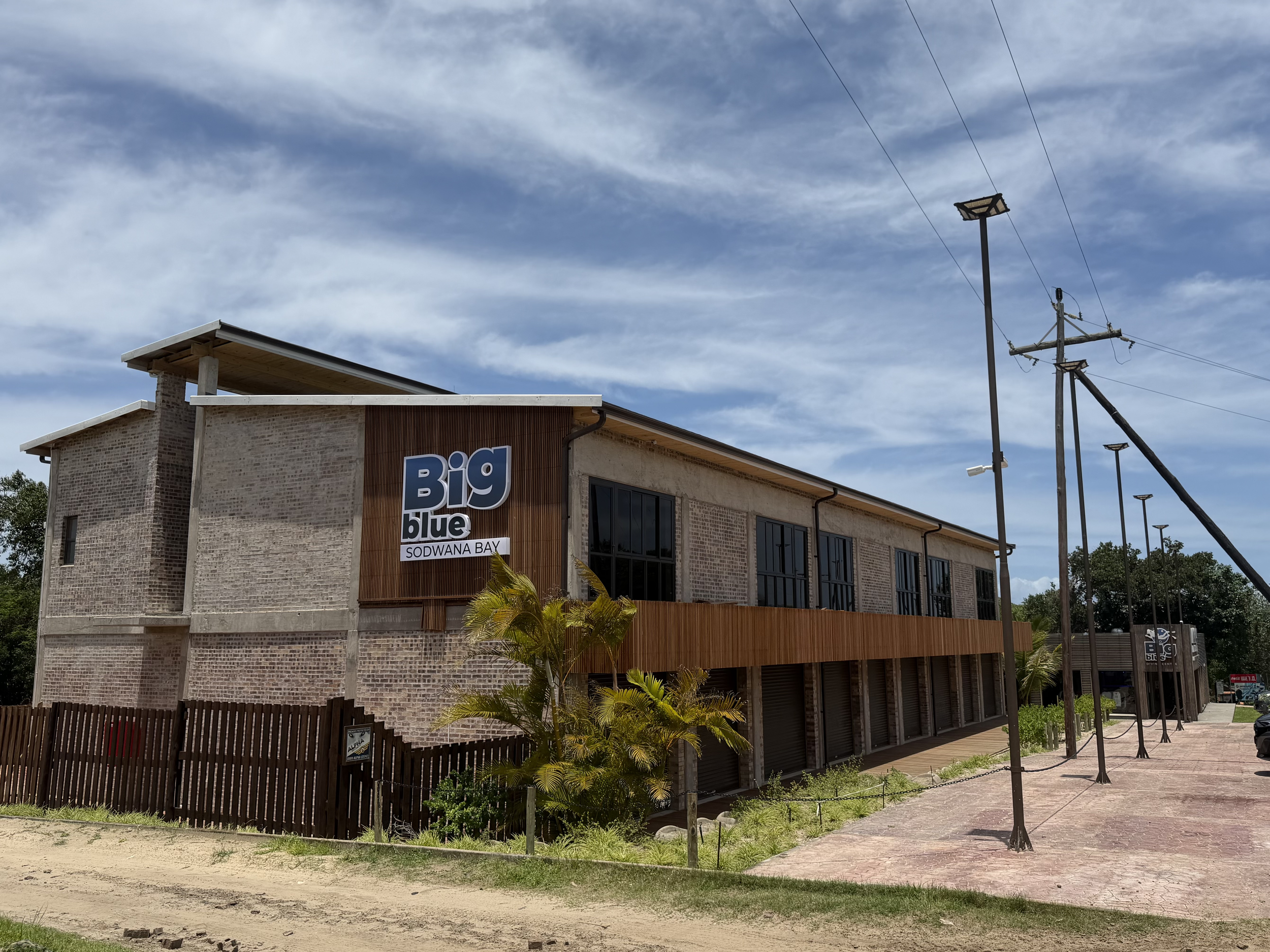 Exterior of a modern brick building with a sign reading "Big blue Sodwana Bay" under a partly cloudy sky. There are utility poles and streetlights nearby, with some greenery and a sidewalk in the foreground.