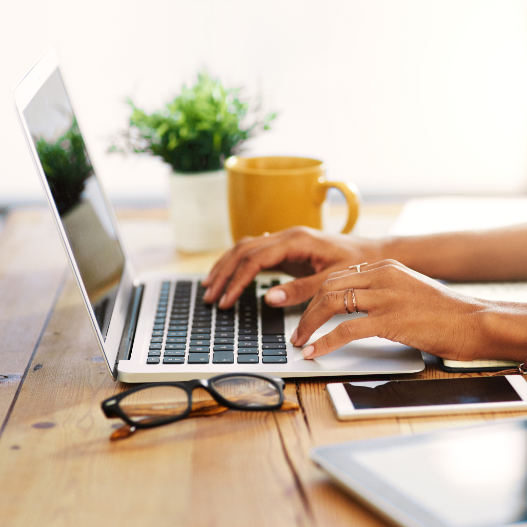 Photo of a woman's hands typing on a laptop with a yellow mug and glasses