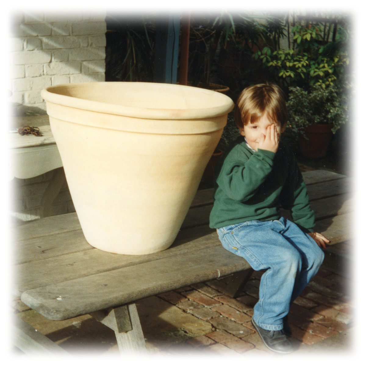 A child in a green sweater sitting on a picnic table next to a large terracotta pot in a garden setting.