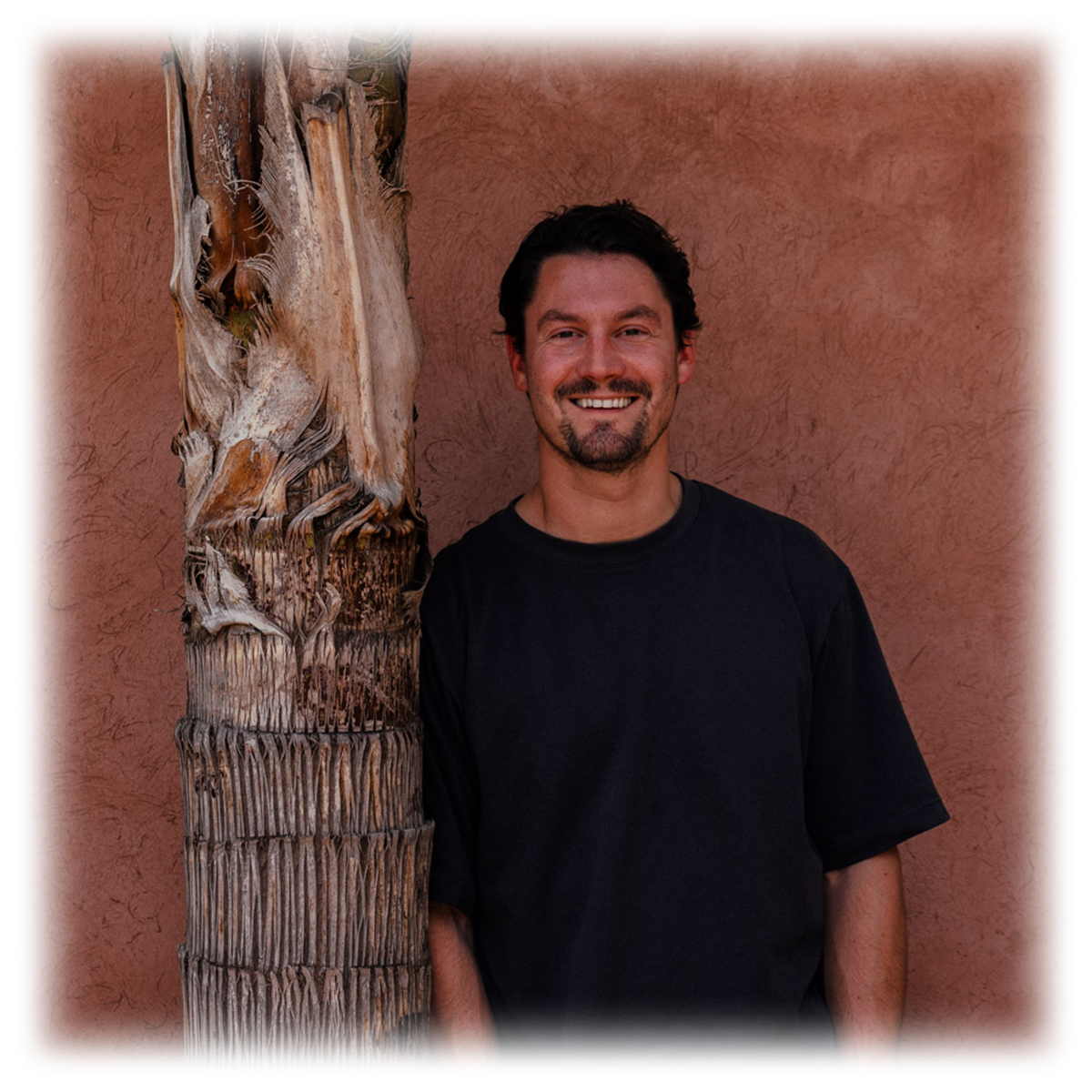 Man in black t-shirt smiling next to palm tree trunk against brown wall