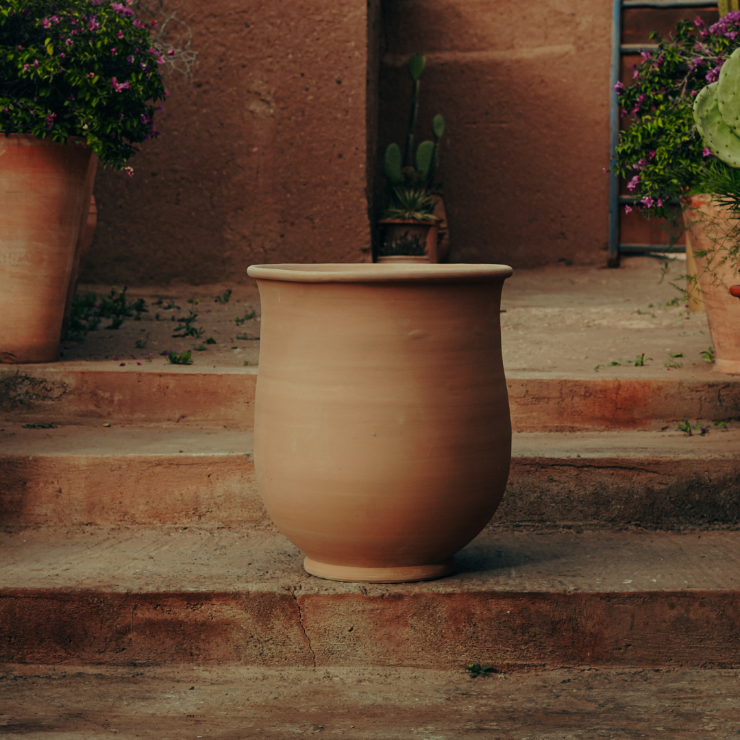 A large, unglazed, natural terracotta pot on outdoor steps, surrounded by potted plants and flowers.