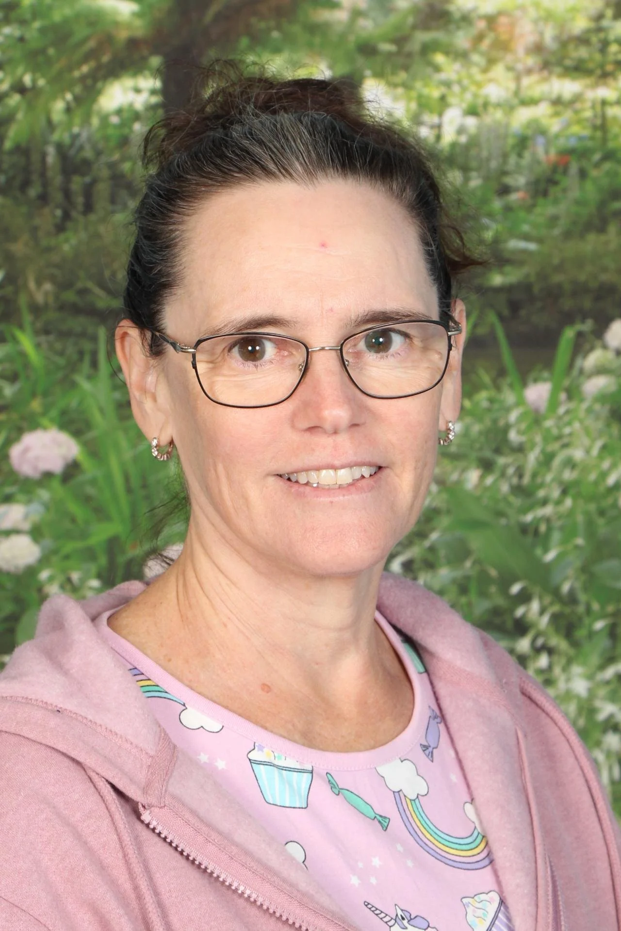 A woman with glasses and earrings, smiling, standing outdoors with greenery and flowers in the background, wearing a pink hoodie and pastel shirt with rainbow, cupcake, and unicorn patterns.