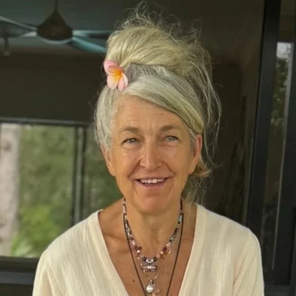 A smiling elderly woman with styled gray hair adorned with a pink flower, wearing layered necklaces and a cream-colored top, standing indoors near a window with greenery outside.