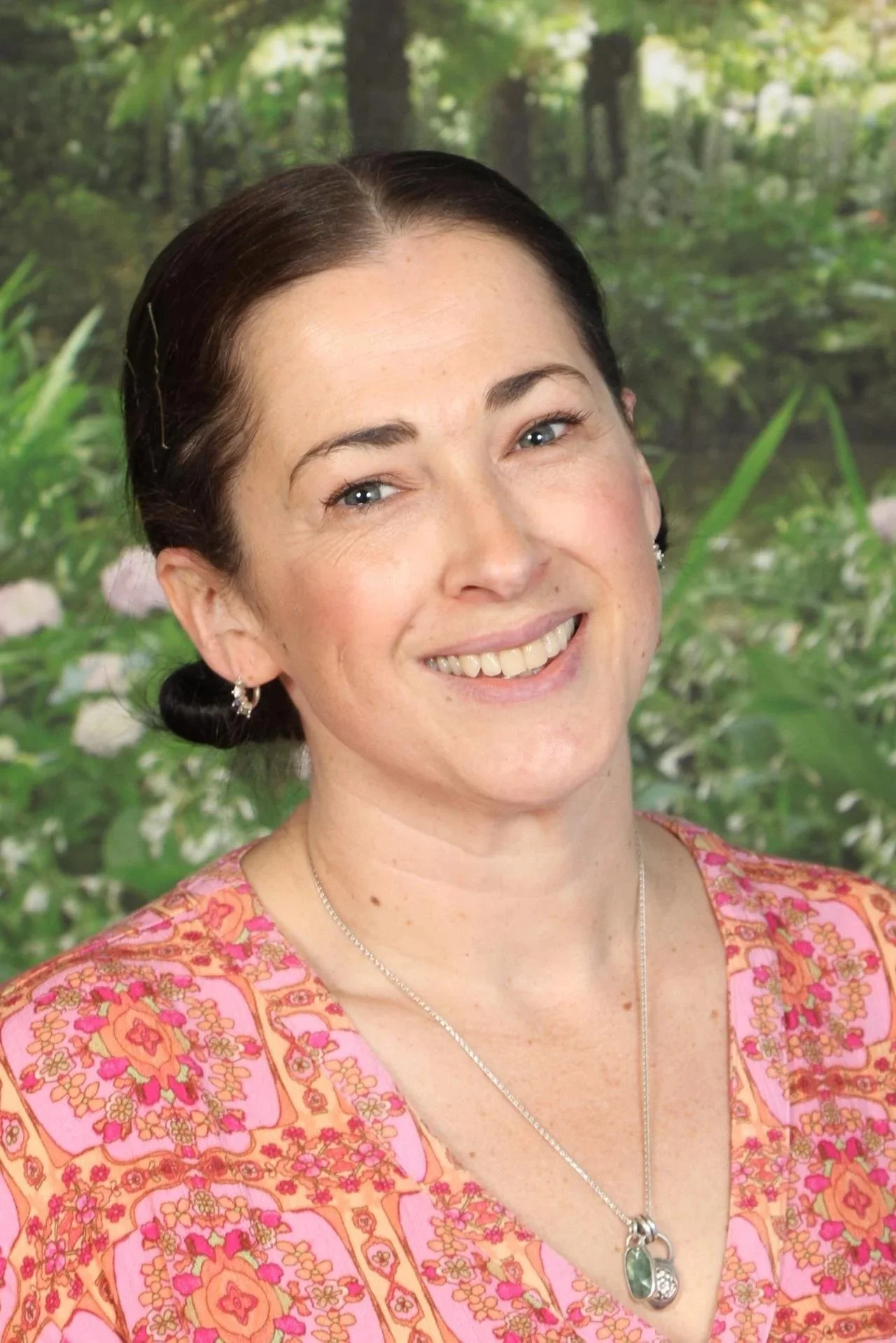 Portrait of a smiling woman with dark hair in a bun, wearing earrings and a necklace with a pendant, in front of a blurred green garden background.