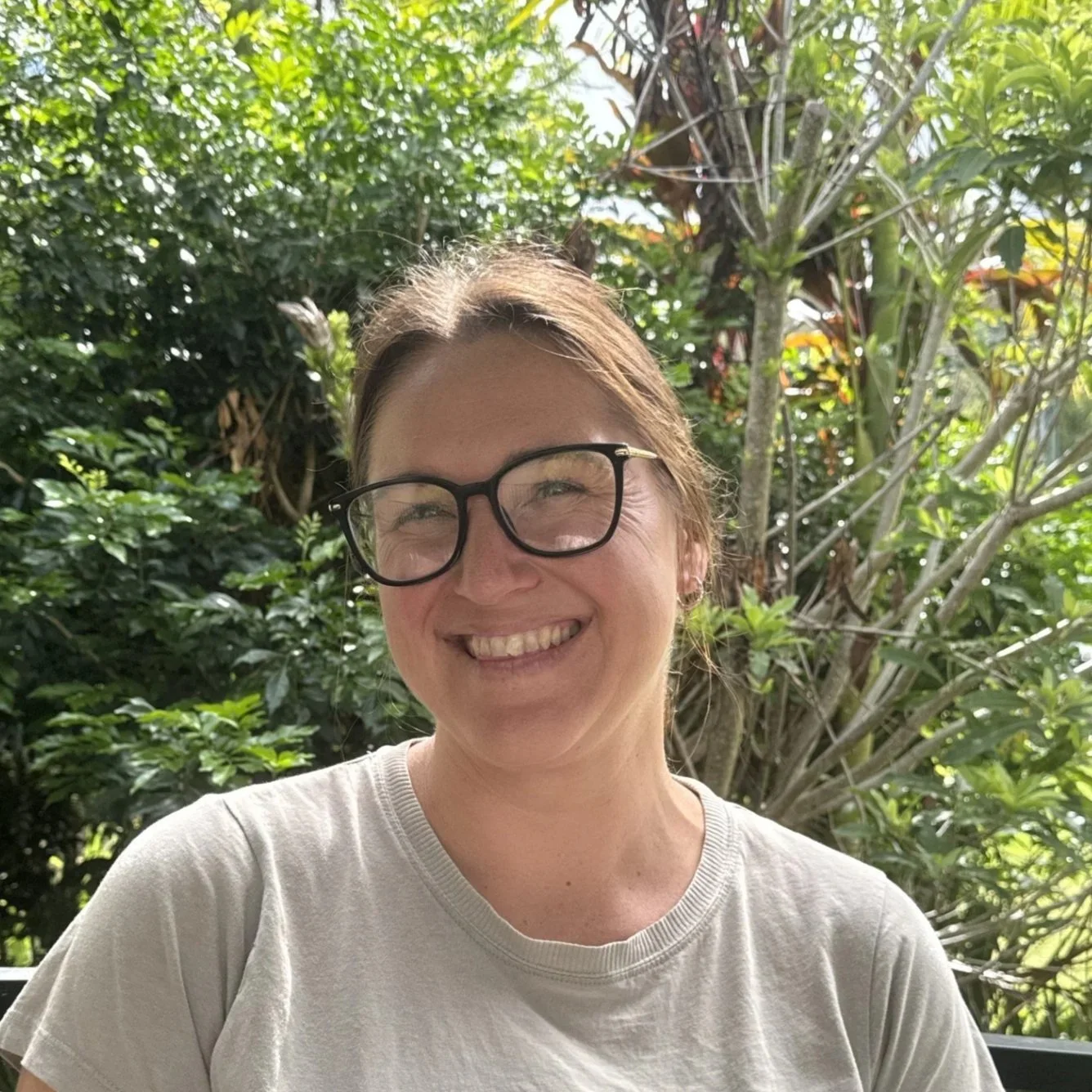 A woman smiling outdoors with green foliage in the background, wearing glasses and a light-colored t-shirt.