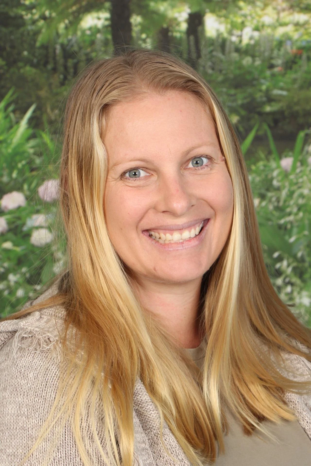 A smiling woman with long blonde hair in front of a background of trees and greenery.