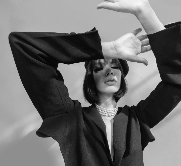 Black and white photo of a stylish person wearing a black jacket, pearl necklace, and beret, posing with arms raised.