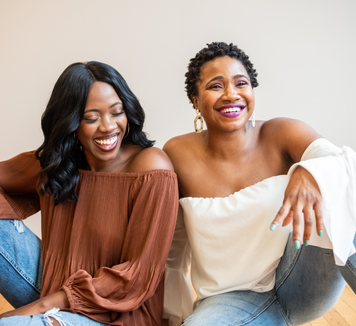 Two women smiling and sitting together, wearing casual off-the-shoulder tops, with a light neutral background.