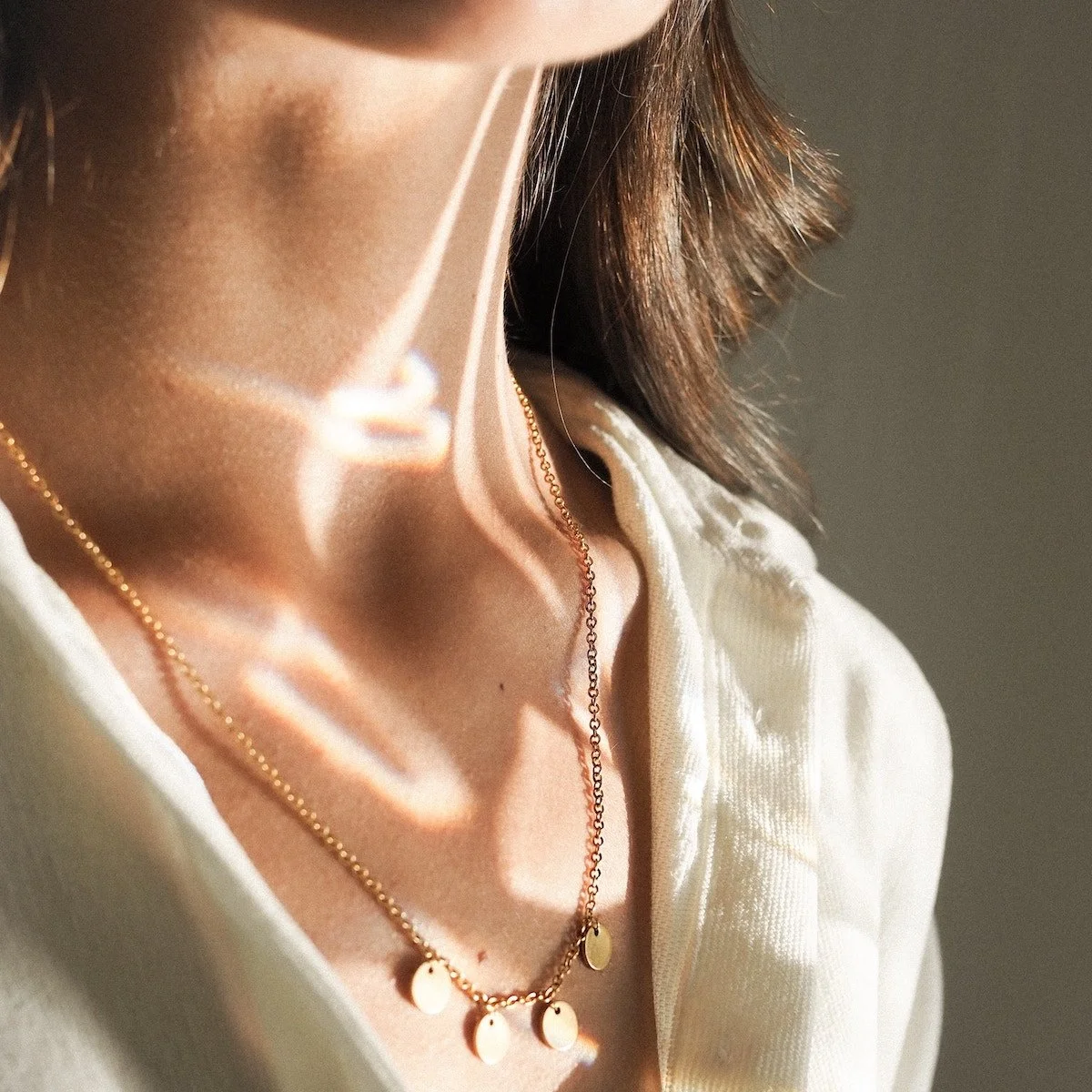 Close-up of a woman's neck wearing a gold disc necklace and a light blouse with sunlight reflections on her skin.