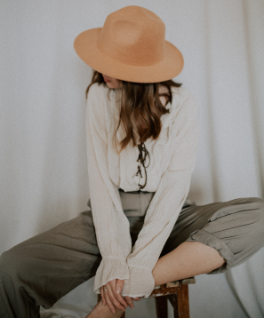 Person wearing a beige hat, white blouse, and gray pants sitting on a stool.