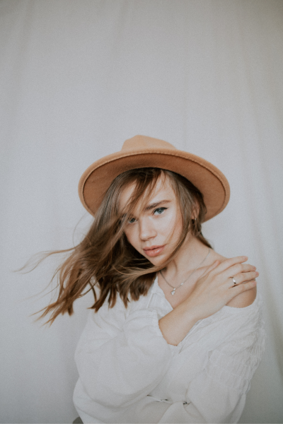 Person wearing a beige hat and white blouse, with windblown hair, posing against a neutral background.