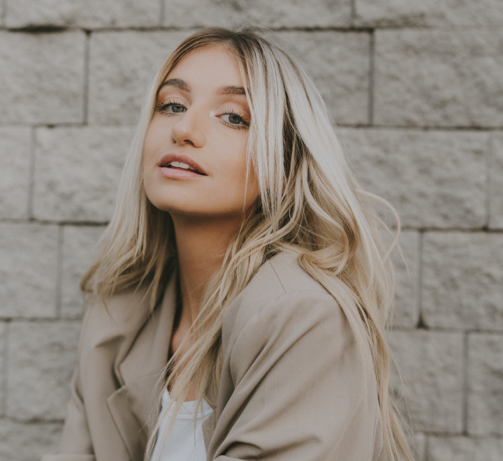 Woman with long blonde hair posing against a brick wall, wearing a beige blazer.