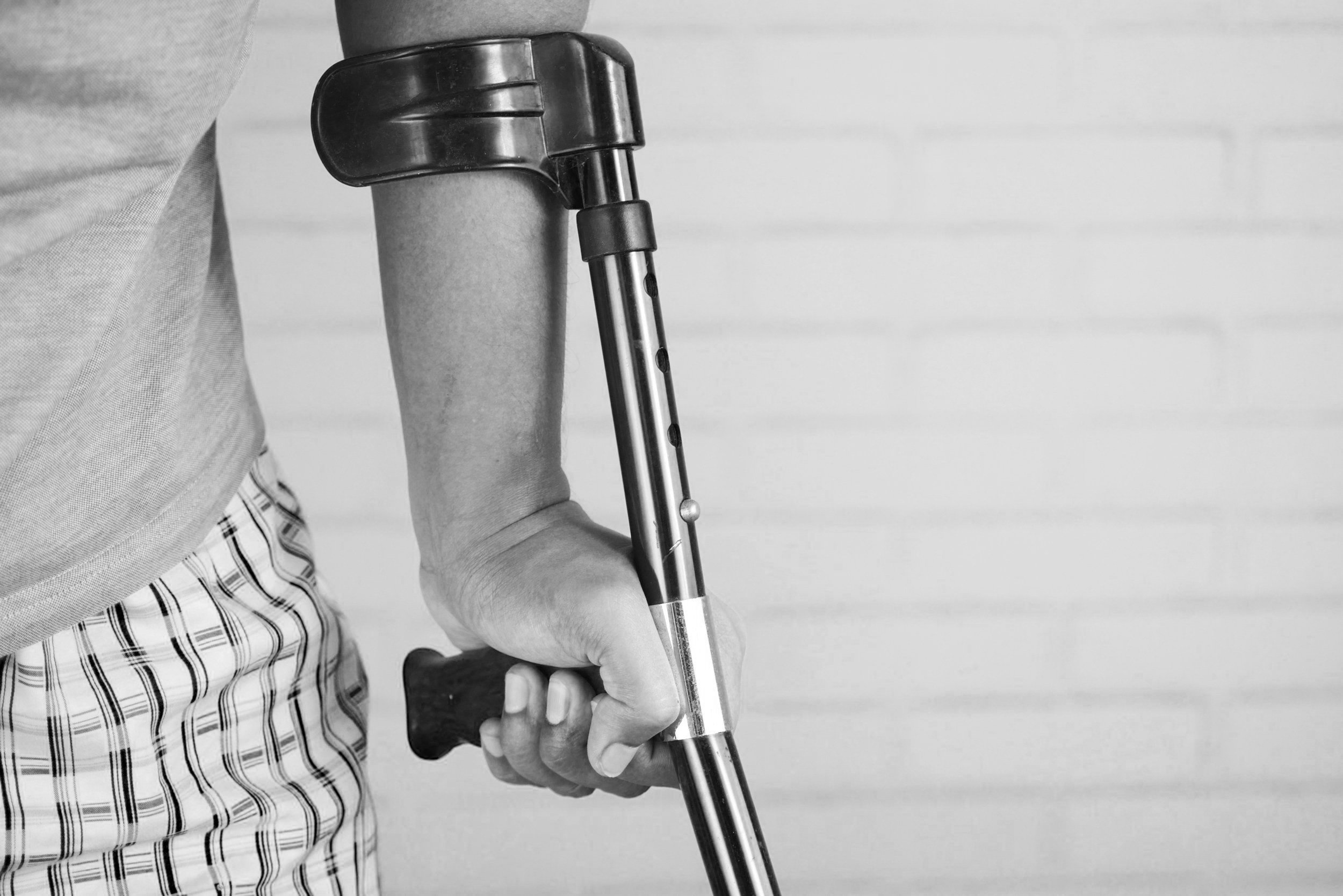 Person using a crutch, wearing plaid pants and a light shirt, against a brick wall background.
